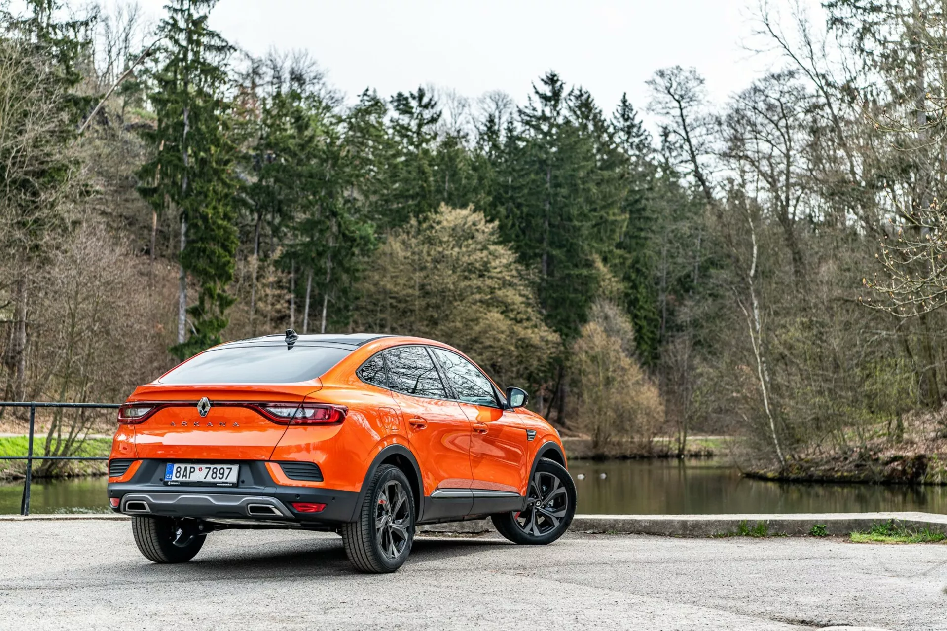 Renault Arkana 2021 visto desde un ángulo trasero de tres cuartos, vista detallada de los perfiles lateral y trasero, mostrando un moderno crossover/SUV con mayor altura del suelo y una deportiva línea de techo estilo cupé. La carrocería naranja brillante destaca contra los árboles y el lago, resaltando las llantas de aleación de diseño multirradio, luces traseras LED, alerón trasero, antena de aleta de tiburón, emblema centralmente ubicado, salidas de escape dobles, acabado de pintura brillante y cristales traseros tintados. La iluminación natural difusa mejora la visibilidad del exterior bien mantenido, sugiriendo un atractivo para consumidores con estilos de vida activos, aparcado sobre una superficie pavimentada.