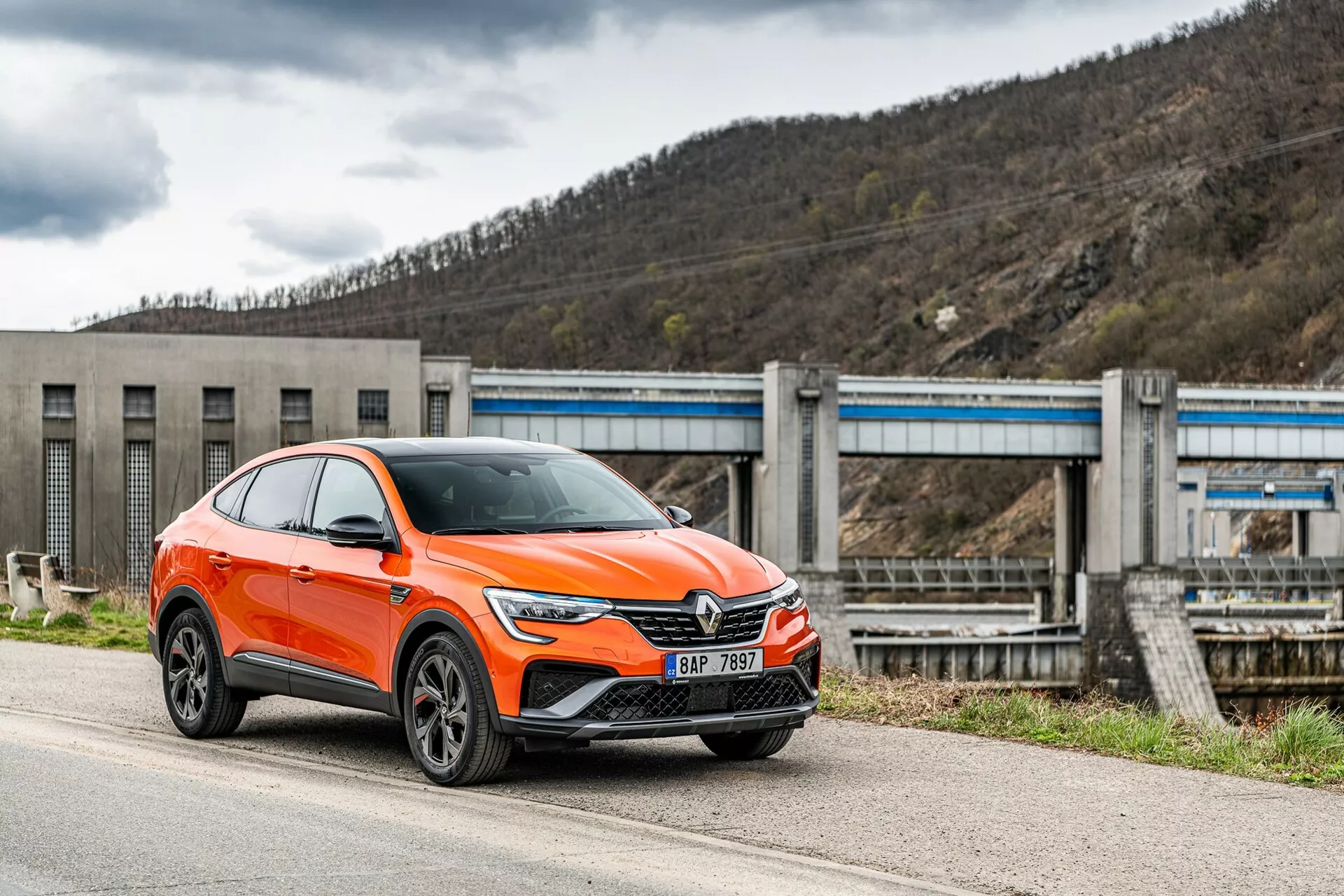 Un moderno Renault Arkana 2021 de color naranja brillante, crossover SUV, está estacionado junto a una carretera, fotografiado desde un ángulo frontal y lateral centrado en el lado del conductor. La imagen muestra la parrilla delantera del auto con un gran emblema, faros LED angulares y elegantes, luces diurnas, entradas de aire y posibles sensores de asistencia al conductor en el parachoques inferior, arcos de guardabarros pronunciados con ruedas de aleación de múltiples radios y espejos con indicadores de giro integrados. El vehículo, clasificado como SUV, tiene una postura elevada con mayor distancia al suelo y un techo inclinado hacia atrás al estilo de un cupé. En el fondo, se puede ver una infraestructura que se asemeja a una presa o un sistema de compuertas, situando el automóvil en un entorno industrial o rural. La toma se realiza desde un ángulo ligeramente bajo, enfatizando la altura y la postura del automóvil.