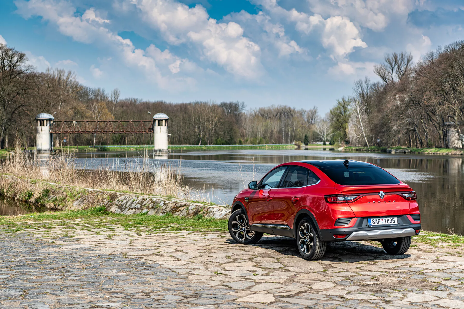 Renault Arkana 2021 estacionado en adoquines cerca del agua con fondo natural, mostrando vista trasera de tres cuartos, diseño deportivo rojo y luces traseras LED