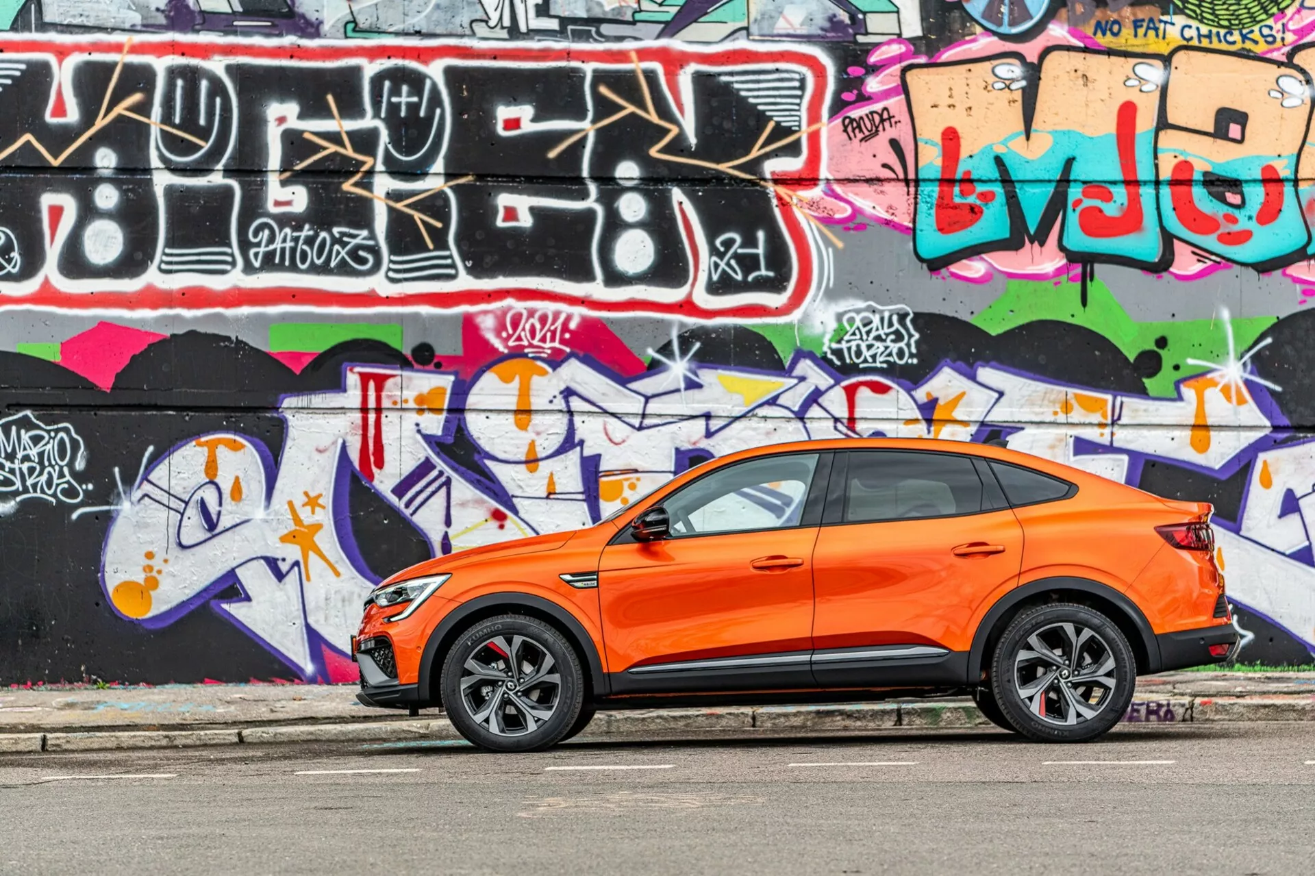La imagen presenta un crossover SUV Renault Arkana 2021 de color naranja vibrante estacionado en paralelo a una calle con una pared de graffiti de colores brillantes en el fondo. Capturada desde un ángulo de vista lateral, la fotografía resalta el diseño deportivo y moderno del vehículo con las puertas delanteras y traseras del lado del conductor visibles, ventanas laterales, espejos laterales y una línea de techo inclinada. Las ruedas de aleación estilizadas con neumáticos de perfil bajo sugieren un enfoque en el rendimiento en carretera. Las manijas de entrada sin llave en las puertas delanteras dan pistas sobre un sistema de entrada y arranque sin llave. La imagen se toma a la luz del día, enfatizando el contraste del cuerpo naranja contra el graffiti, mostrando la carrocería bien mantenida y nítida del automóvil en un entorno urbano y juvenil.