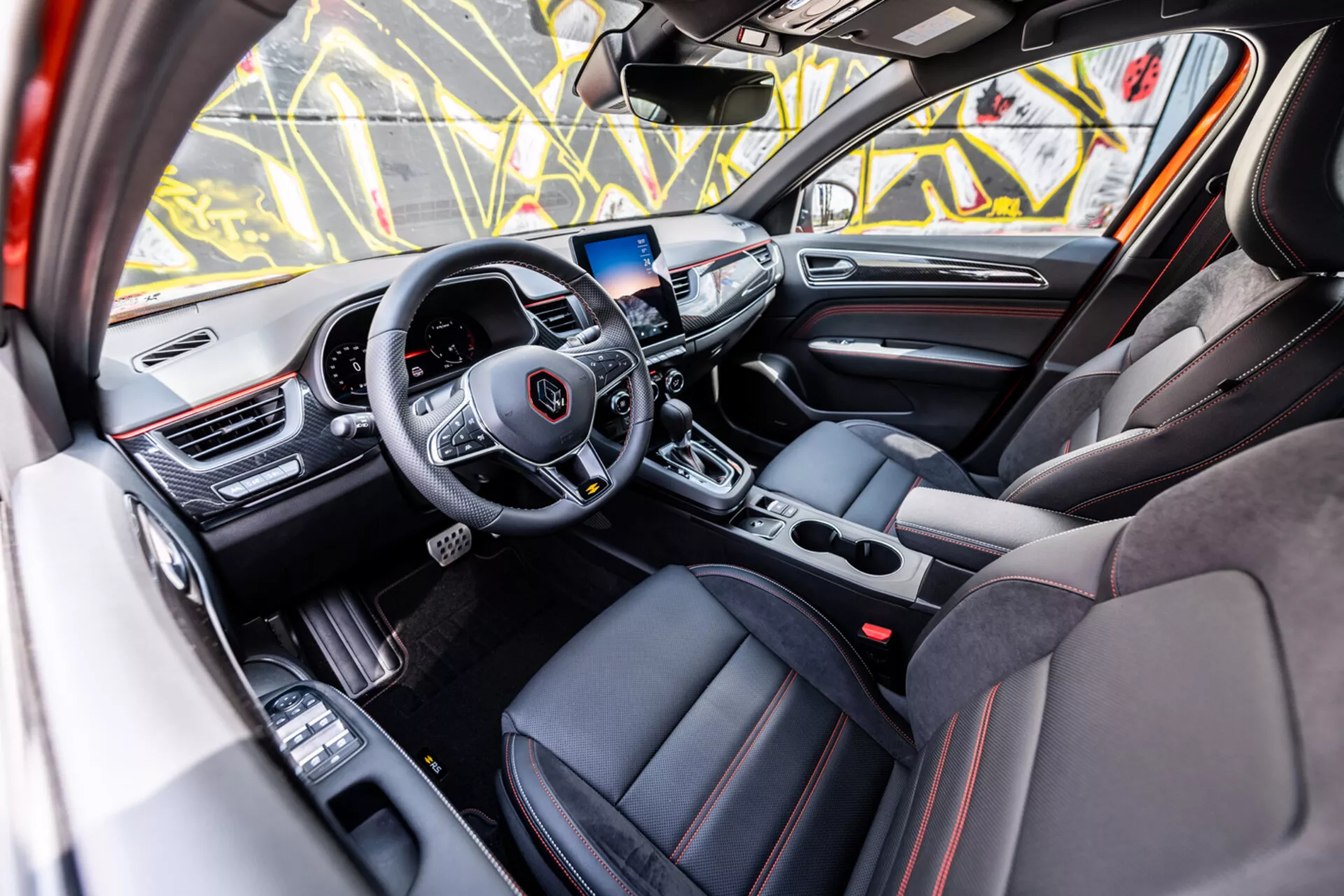 Interior del Renault Arkana 2021 mostrando la lujosa cabina delantera desde la perspectiva de la puerta del pasajero con un volante moderno de tres radios, pantalla táctil de entretenimiento y tapicería deportiva con costuras rojas