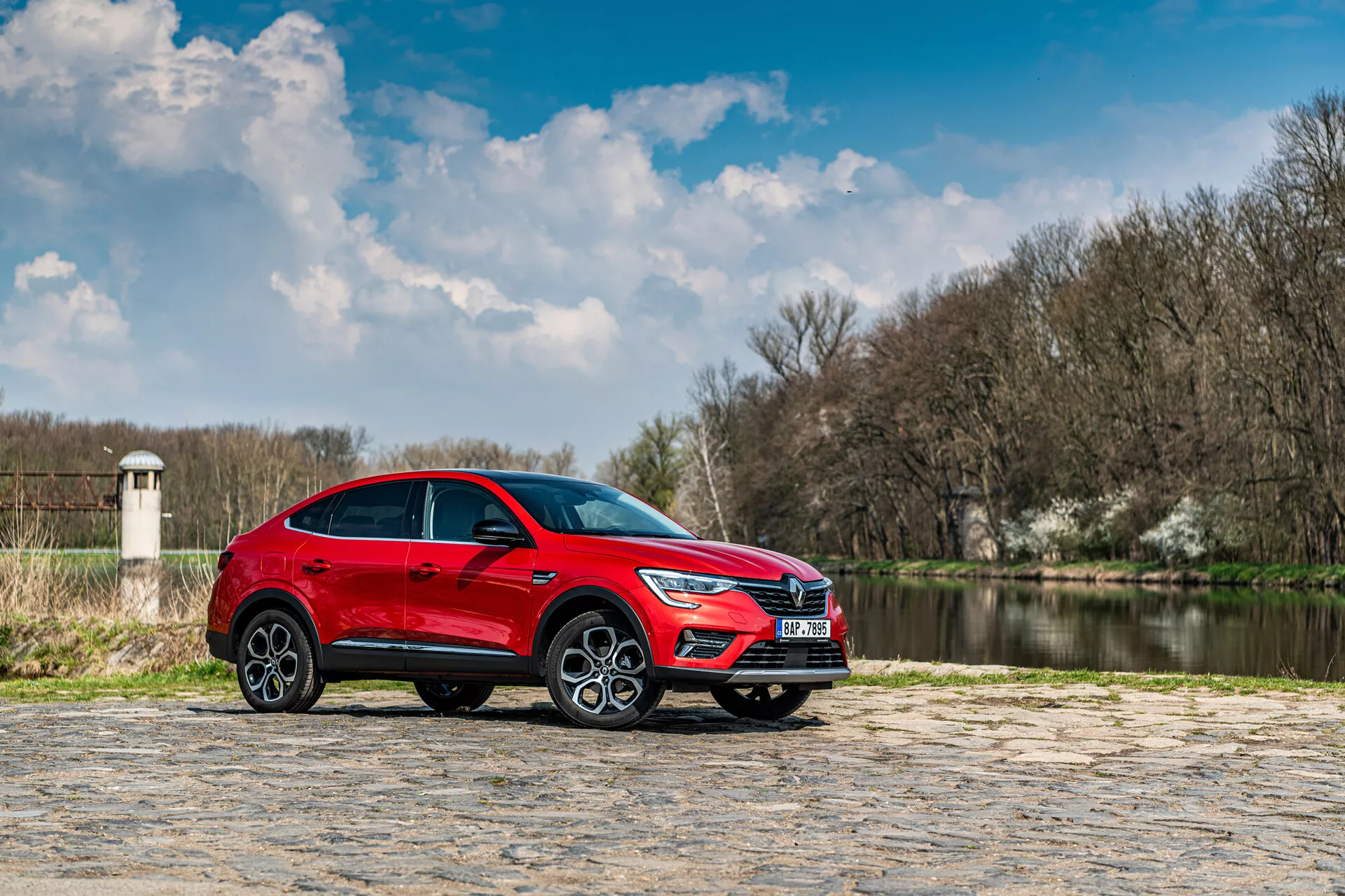 SUV rojo Renault Arkana 2021 estacionado en pavimento de piedra con cuerpo de agua y árboles al fondo bajo un cielo parcialmente nublado, perfil lateral destaca su diseño elegante y características dinámicas