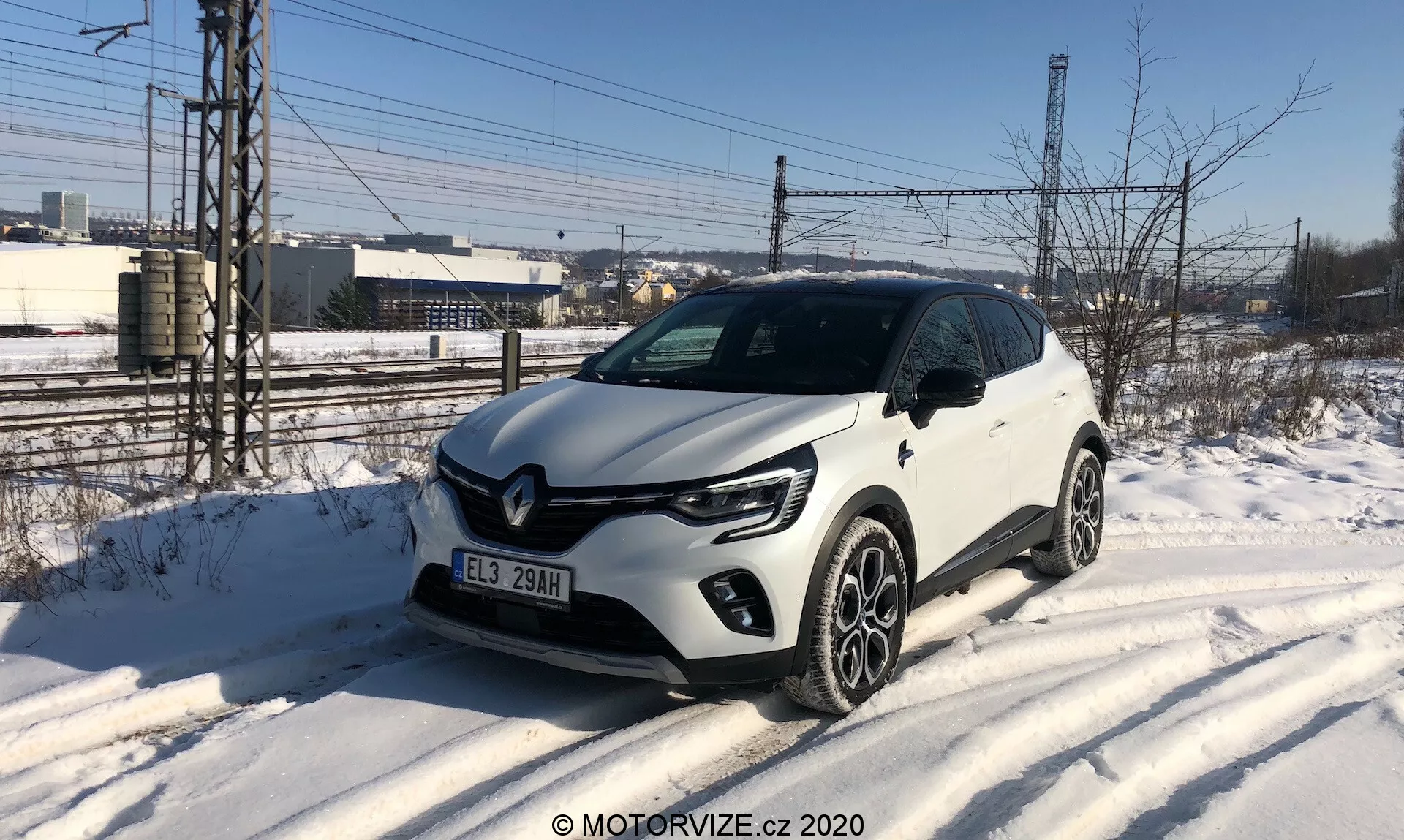 La imagen muestra un Renault Captur 2020 blanco, un SUV compacto estacionado en un borde de carretera cubierto de nieve, capturado desde un ángulo frontal de tres cuartos. El vehículo presenta luces diurnas LED en forma de C, faros proyector, una parrilla frontal con acentos negros y una insignia Renault prominente, y llantas de aleación de varios radios. Elementos de diseño notables incluyen el revestimiento de plástico en los bordes inferiores y los arcos de las ruedas. El ambiente sugiere un día frío de invierno con un fondo industrial o semiurbano.