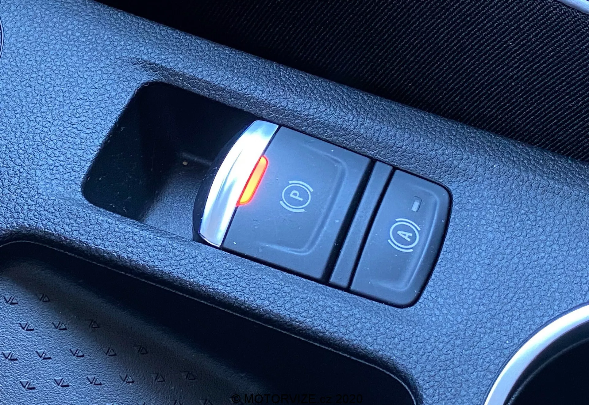 Plano diagonal de cerca del interruptor del freno de mano electrónico en el interior del Renault Captur 2020. El interruptor, ubicado en la consola central, presenta dos botones: uno rojo iluminado con un candado cerrado y el símbolo de 'P' indicando el estado activado, y otro botón con el símbolo de candado abierto. El interruptor está colocado en una consola de plástico texturizado negro, destacando la tecnología moderna del vehículo.