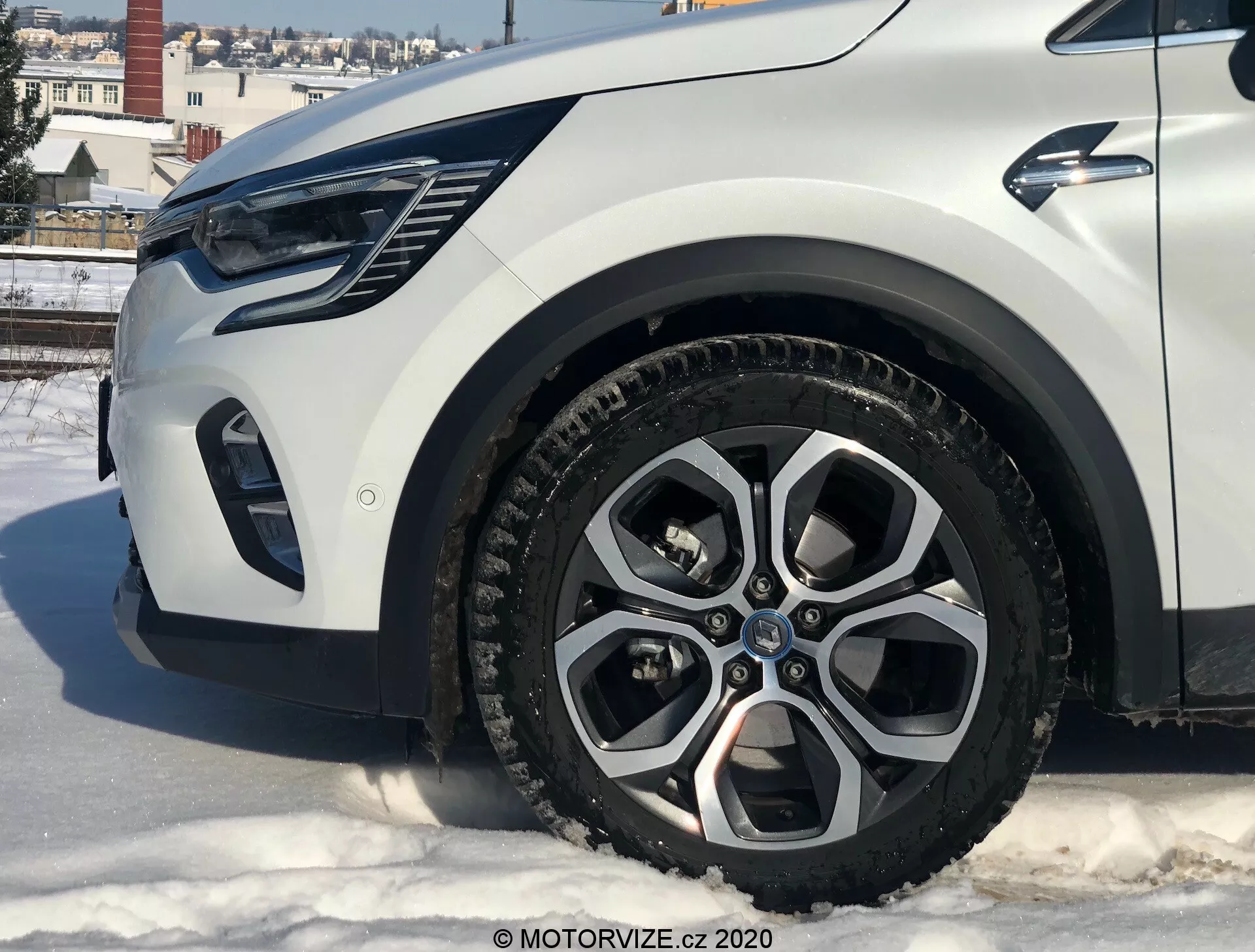 Primer plano de un Renault Captur 2020 desde un ángulo bajo que muestra la sección delantera izquierda, con enfoque en la rueda de aleación, el guardabarros, el parachoques y el área de los faros. El coche está sobre una superficie nevada en un entorno urbano con edificios y cielo despejado.
