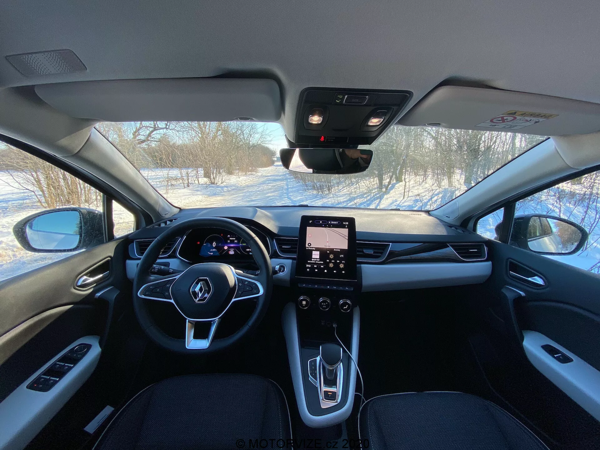 Toma del interior del Renault Captur 2020 desde los asientos traseros mostrando la cabina delantera con volante de tres radios con botones integrados, panel de instrumentos digital, sistema de infoentretenimiento con pantalla táctil verticalmente orientada, consola central moderna con pocos controles físicos, selector de marchas de diseño elegante, ventilaciones de HVAC, materiales premium incluidos cuero y acentos metálicos, espejo retrovisor posiblemente con sensores para ADAS y pilar A visible con controles de ventanas.
