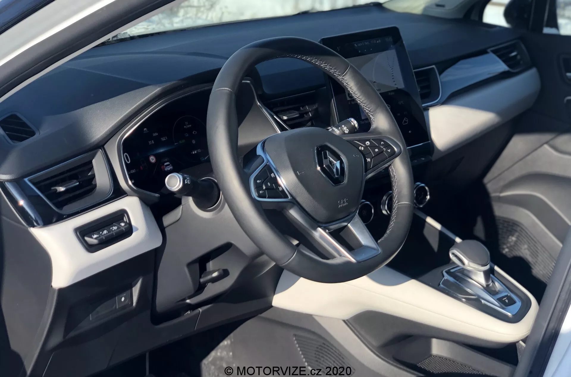 Vista interior del Renault Captur 2020 desde la perspectiva del conductor, que muestra el volante con el logo de Renault, controles montados, panel de instrumentos digital detrás y un sistema de infoentretenimiento con pantalla táctil centralmente ubicado. La imagen destaca una mezcla de materiales de cuero sintético y plástico con acentos metálicos, con transmisión automática, sistema de control de clima y ajustes de ventanas y espejos eléctricos.
