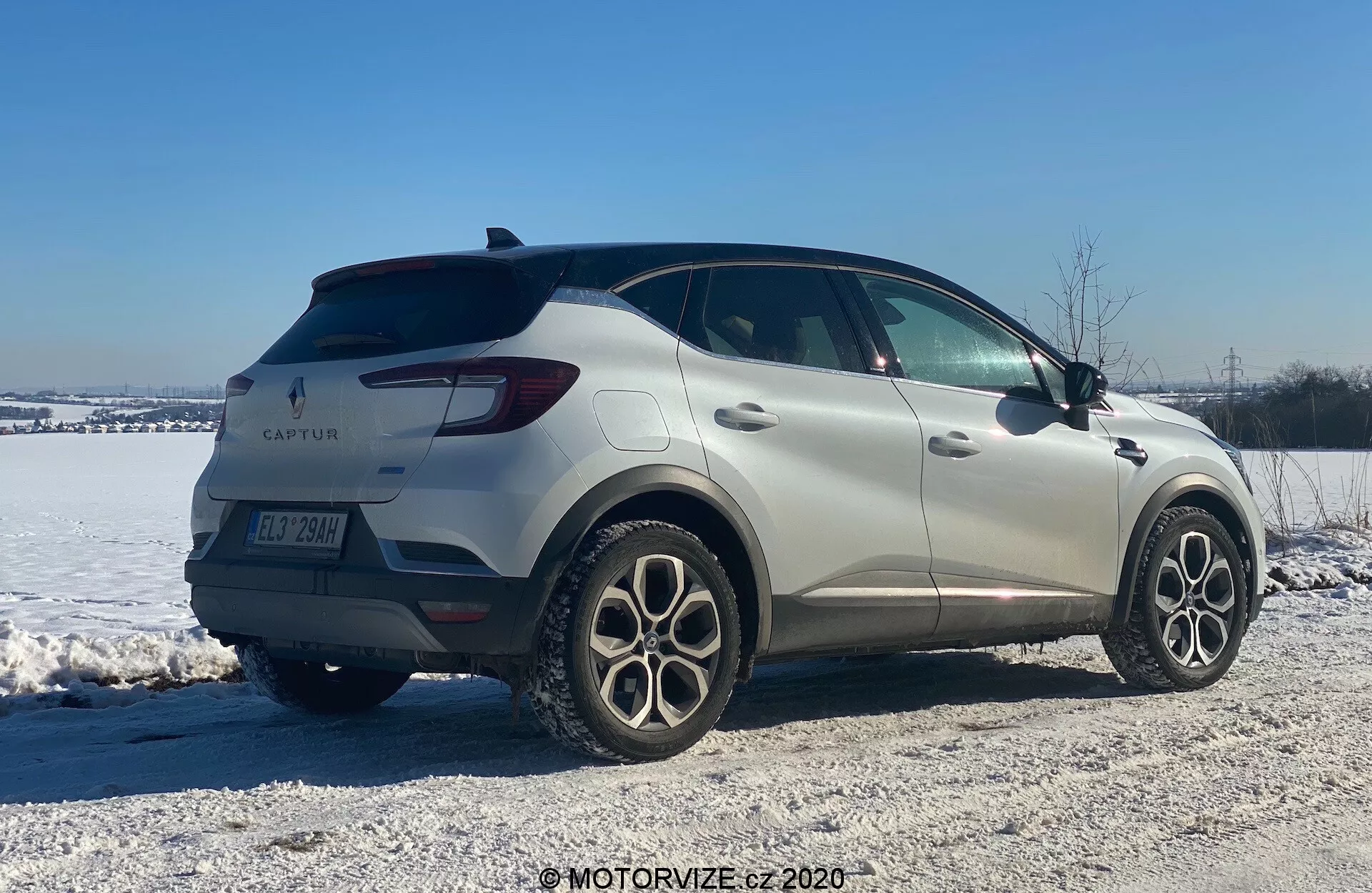Imagen del Renault Captur 2020, un SUV compacto, en un entorno invernal. Mostrado desde una vista trasera de tres cuartos, el coche exhibe un diseño elegante con líneas suaves y luces traseras LED. El emblema del fabricante y el nombre del modelo son visibles junto con llantas de aleación de múltiples radios, sensores de estacionamiento traseros y espejos laterales con señales de giro integradas. Colocado en un área cubierta de nieve y grava, la imagen sugiere destreza en terrenos variados y condiciones invernales.