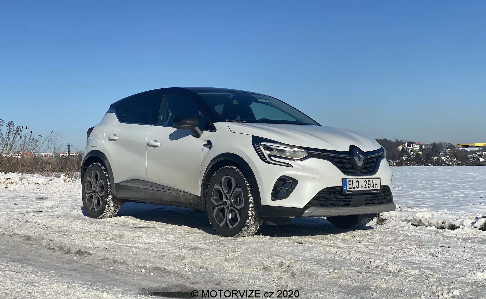Vista de tres cuartos de un SUV crossover compacto Renault Captur 2020 blanco en un escenario invernal, con suelo cubierto de nieve y cielo azul claro. La imagen resalta el lado izquierdo delantero y parte del perfil lateral del vehículo, tomada desde un ángulo ligeramente inferior que enfatiza las ruedas y la mitad inferior. El diseño moderno del coche muestra líneas distintivas en el capó, luces LED elegantes con luces diurnas y una parrilla delantera prominente. Las ruedas de aleación únicas y el revestimiento plástico alrededor de los arcos de las ruedas le añaden un aspecto robusto. Las sombras bajo el coche sugieren que la foto fue tomada a mediodía o por la tarde en luz diurna natural.