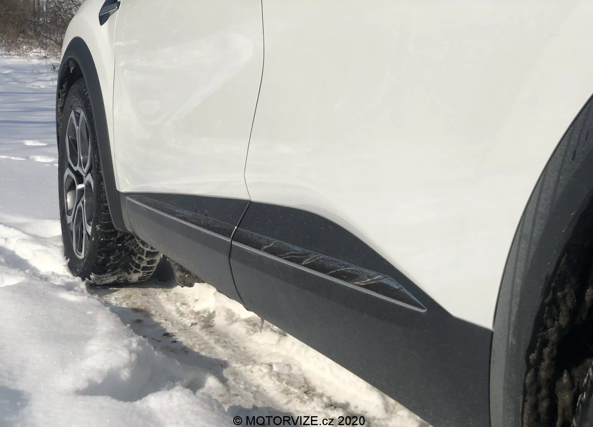 Vista lateral en primer plano y en ángulo desde el nivel del suelo de la rueda delantera y el guardabarros del lado del conductor de un Renault Captur 2020. La llanta de aleación de múltiples radios con neumático de invierno o para todas las estaciones está rodeada de nieve. La carrocería blanca con moldura negra corre a lo largo del borde inferior, indicando un crossover o SUV. La iluminación sugiere un día soleado con sombras visibles.