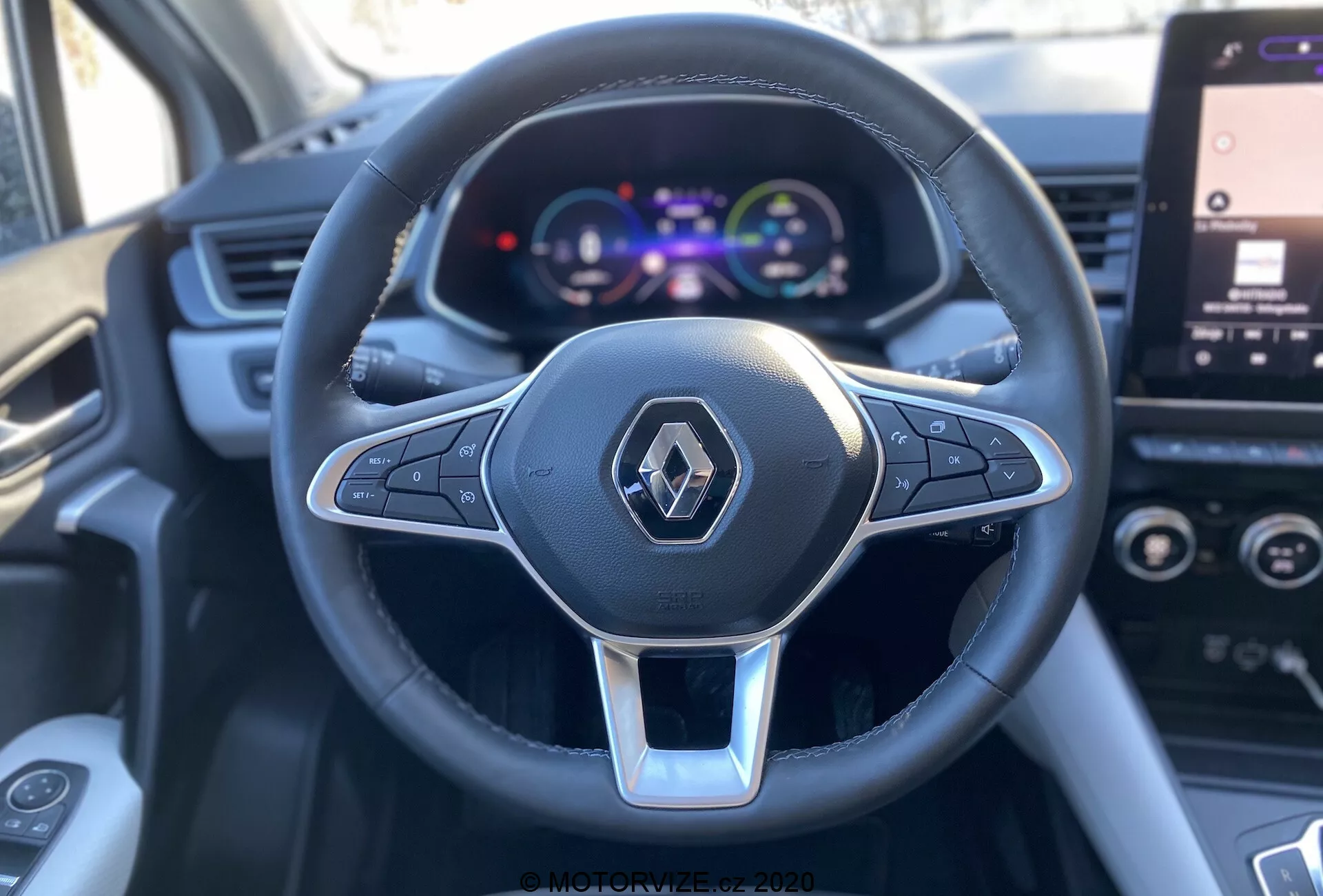 Vista cercana en primera persona desde el asiento del conductor de un Renault Captur 2020, enfocada en el volante multifuncional de tres radios forrado en cuero con acento plateado y logotipo de Renault, junto a un conjunto de instrumentos digital a color que muestra el velocímetro y detalles del vehículo, con una pantalla táctil de infoentretenimiento parcialmente visible en el fondo.