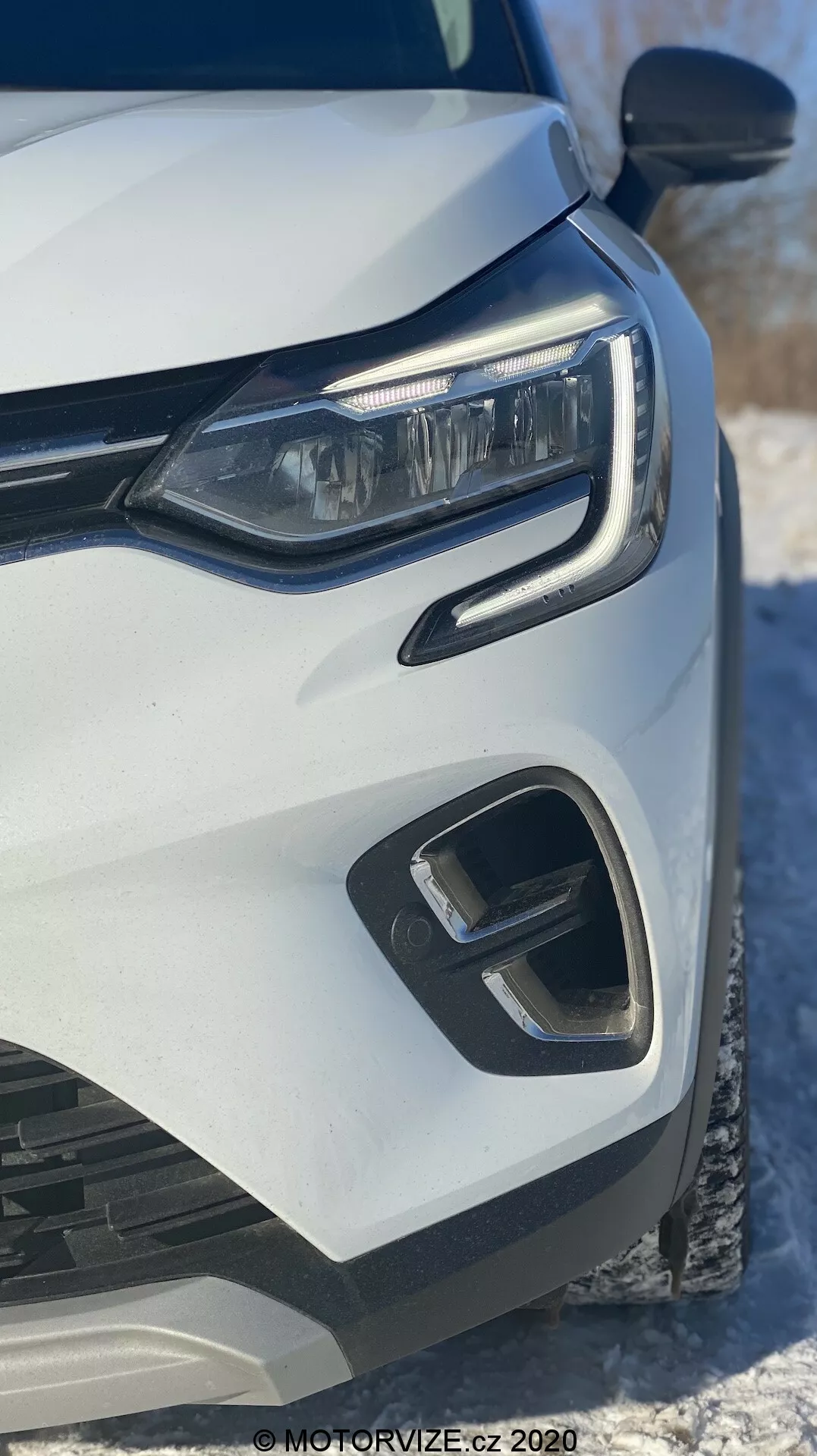 Primer plano del lado izquierdo delantero de un Renault Captur 2020 blanco, que destaca el faro LED moderno, el parachoques delantero geométrico y los elementos de la parrilla, capturado desde un ángulo frontal bajo con nieve en el fondo