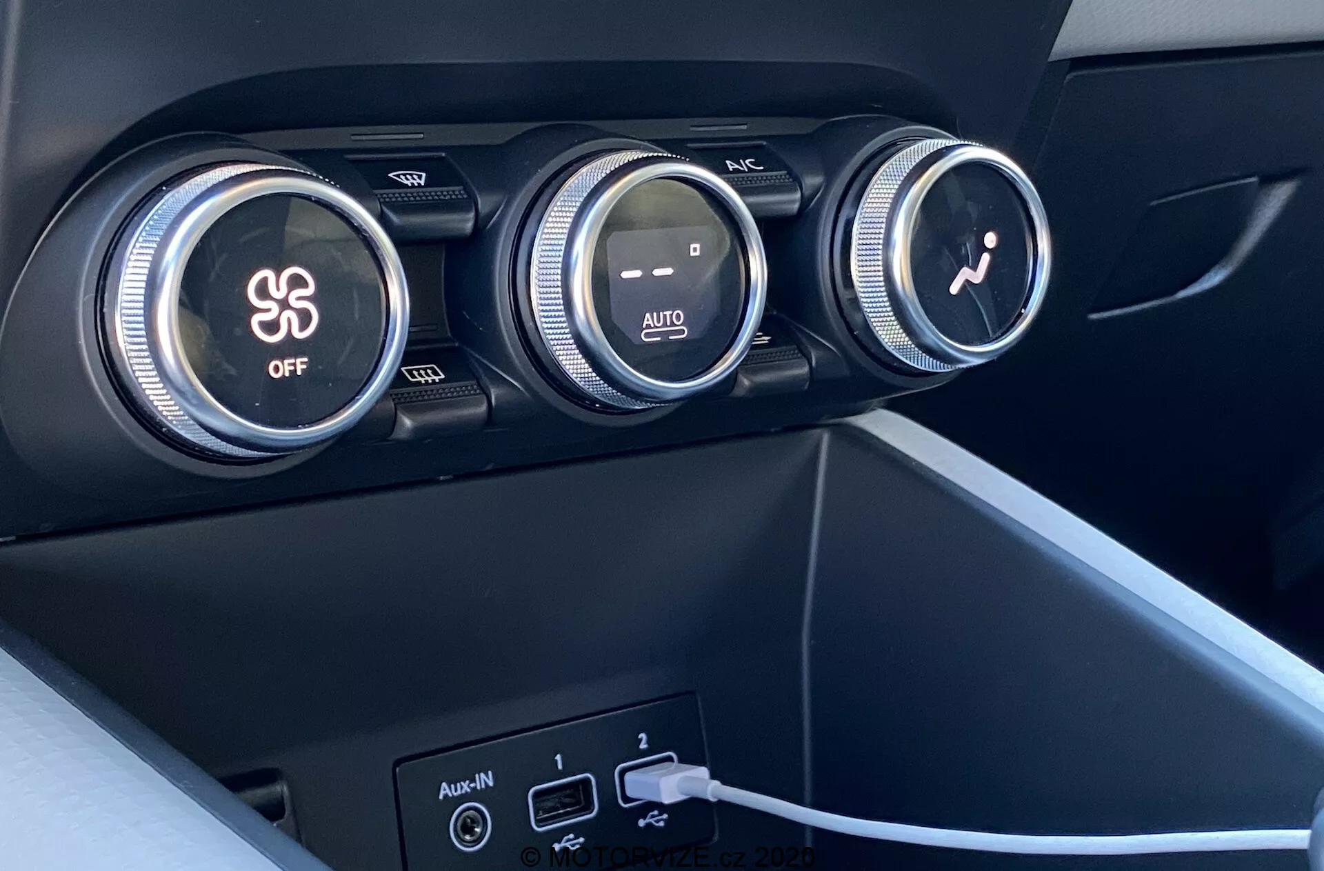 Primer plano del interior del Renault Captur 2020 centrado en el sistema de control de clima. La imagen muestra tres grandes diales en el tablero. El primer dial controla el aire acondicionado, indicado por el símbolo de un copo de nieve y un botón 'OFF'. El dial central regula la temperatura con gradientes de color rojo y azul y una indicación 'AUTO'. El tercer dial con un icono de ventilador ajusta la velocidad del ventilador. Debajo de los diales, hay dos puertos USB y una entrada auxiliar, con un cable conectado a uno de los puertos USB. Los materiales son de plástico oscuro con acabado mate o sintéticos y de diseño moderno y minimalista.
