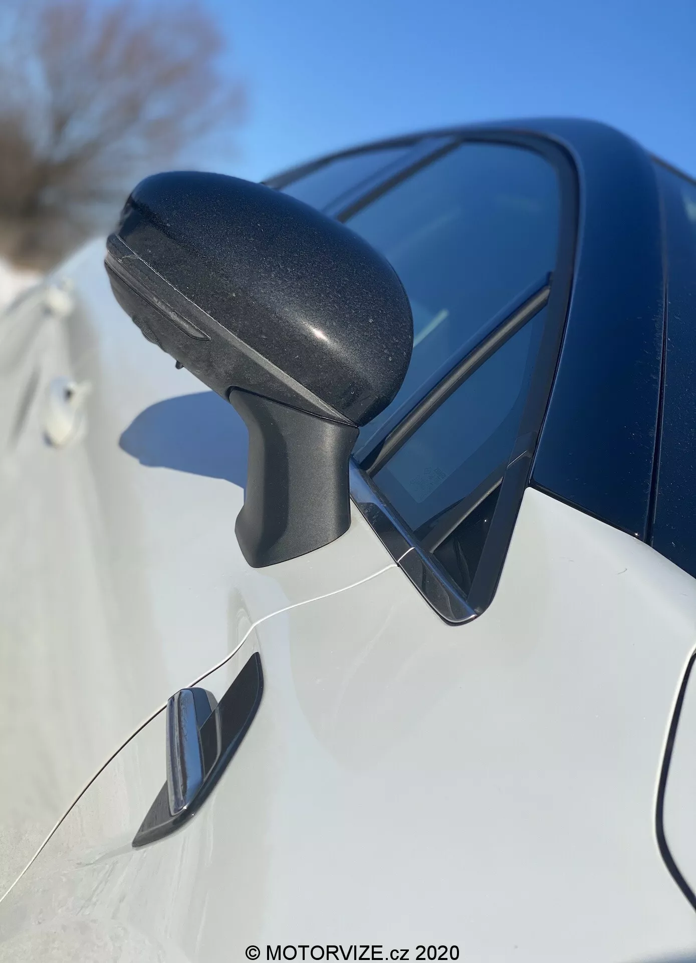 Vista de primer plano del conjunto del espejo lateral del Renault Captur 2020 de adelante hacia atrás, enfocándose en la carcasa de plástico texturizado negro del espejo con una señal de giro integrada y reflejando el cielo azul. El elegante panel del cuerpo de color oscuro debajo y el posible espejo de punto ciego indican un diseño moderno y características avanzadas.