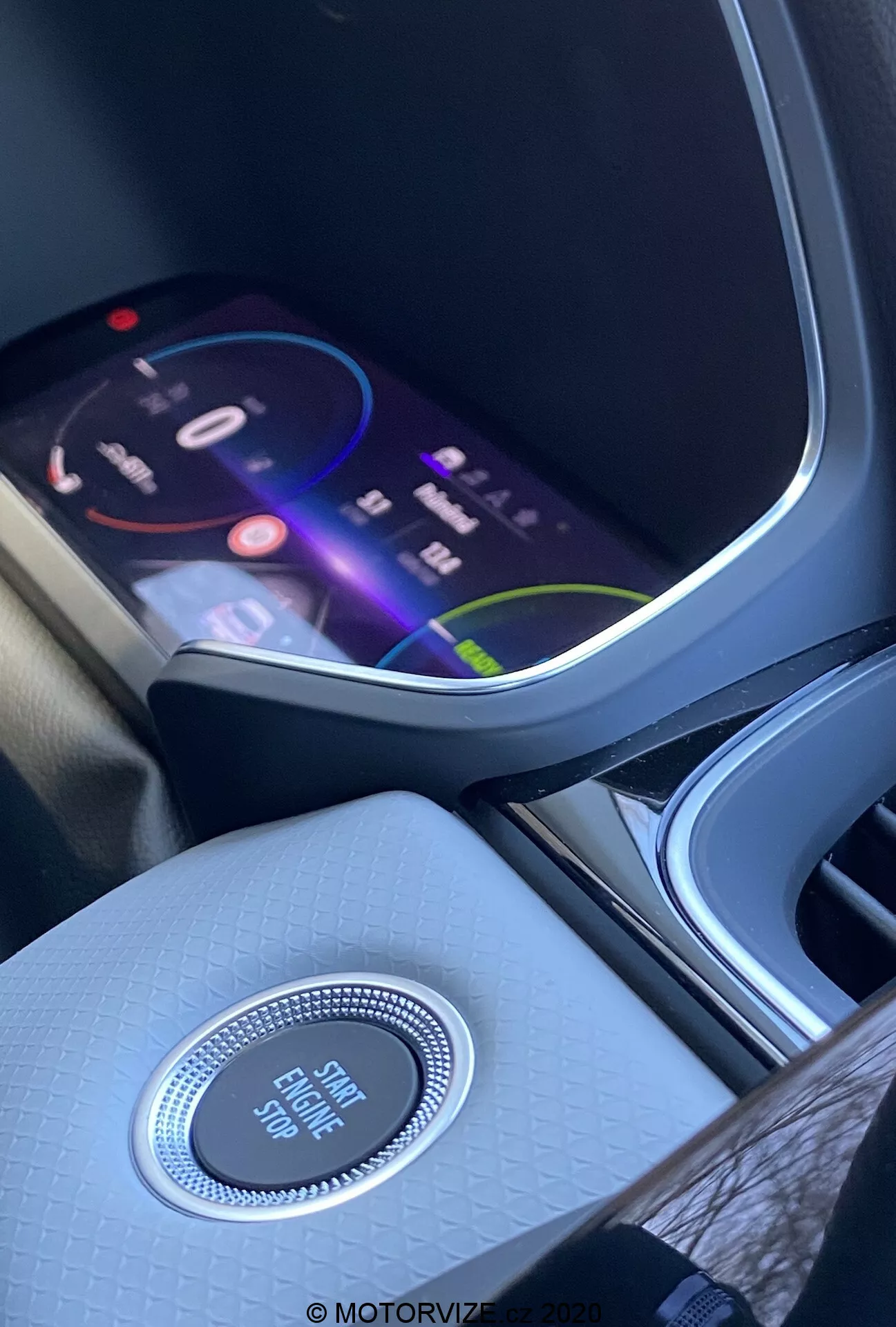 Interior del Renault Captur 2020 enfocándose en la consola central y el tablero desde una perspectiva del asiento delantero. La pantalla de infoentretenimiento vibrante muestra una interfaz colorida que sugiere características multimedia avanzadas, incluyendo navegación, reproducción multimedia y opciones de conectividad. Debajo de la pantalla, un control circular con acabado cromado etiquetado como 'Engine Start Stop' destaca el sistema de arranque sin llave. La consola central tiene un acabado texturizado de alta calidad, mejorando la ambientación interior. A la izquierda, se puede ver parcialmente un portavasos o compartimento de almacenamiento.