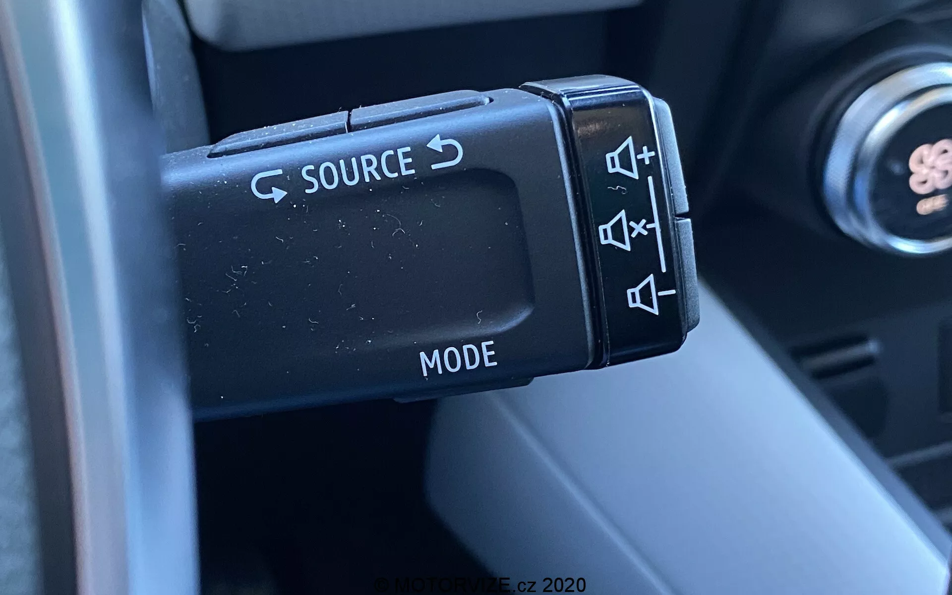 Primer plano de los controles de audio montados en el volante de un Renault Captur 2020, centrándose en la palanca de control del lado izquierdo con botones para cambiar la fuente (SOURCE), ajuste de volumen, salto de pista y selección de modo (MODE). La fotografía captura los detalles de los botones de plástico con acabado mate y su diseño ergonómico, con parte de la columna de dirección y el interior desenfocados en el fondo, resaltando la funcionalidad y la conveniencia para el conductor.