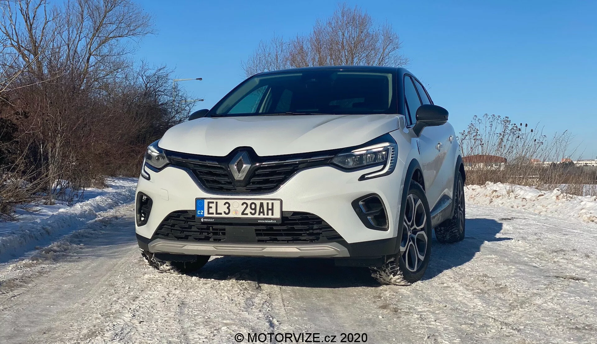 Vista frontal de un Renault Captur blanco 2020, fotografiado en un ángulo que favorece ligeramente el lado izquierdo. El vehículo moderno exhibe un distintivo diseño de parrilla frontal Renault con un gran logo central, líneas angulares y contornos esculpidos. Se muestran claramente la parrilla frontal completa, los faros LED y las luces diurnas angulares. El parachoques deportivo tiene entradas de aire laterales prominentes y una sección central probablemente para enfriamiento. El capó tiene líneas elevadas para una apariencia musculosa. Parte de las ruedas delanteras y las llantas de aleación de múltiples radios son visibles en la superficie nevada, indicando condiciones invernales. La matrícula europea y la pintura brillante que refleja la luz solar sugieren un día soleado y claro. Sistemas avanzados de asistencia al conductor, iluminación LED y tren motriz híbrido/eléctrico inferidos del 'E' en la matrícula.