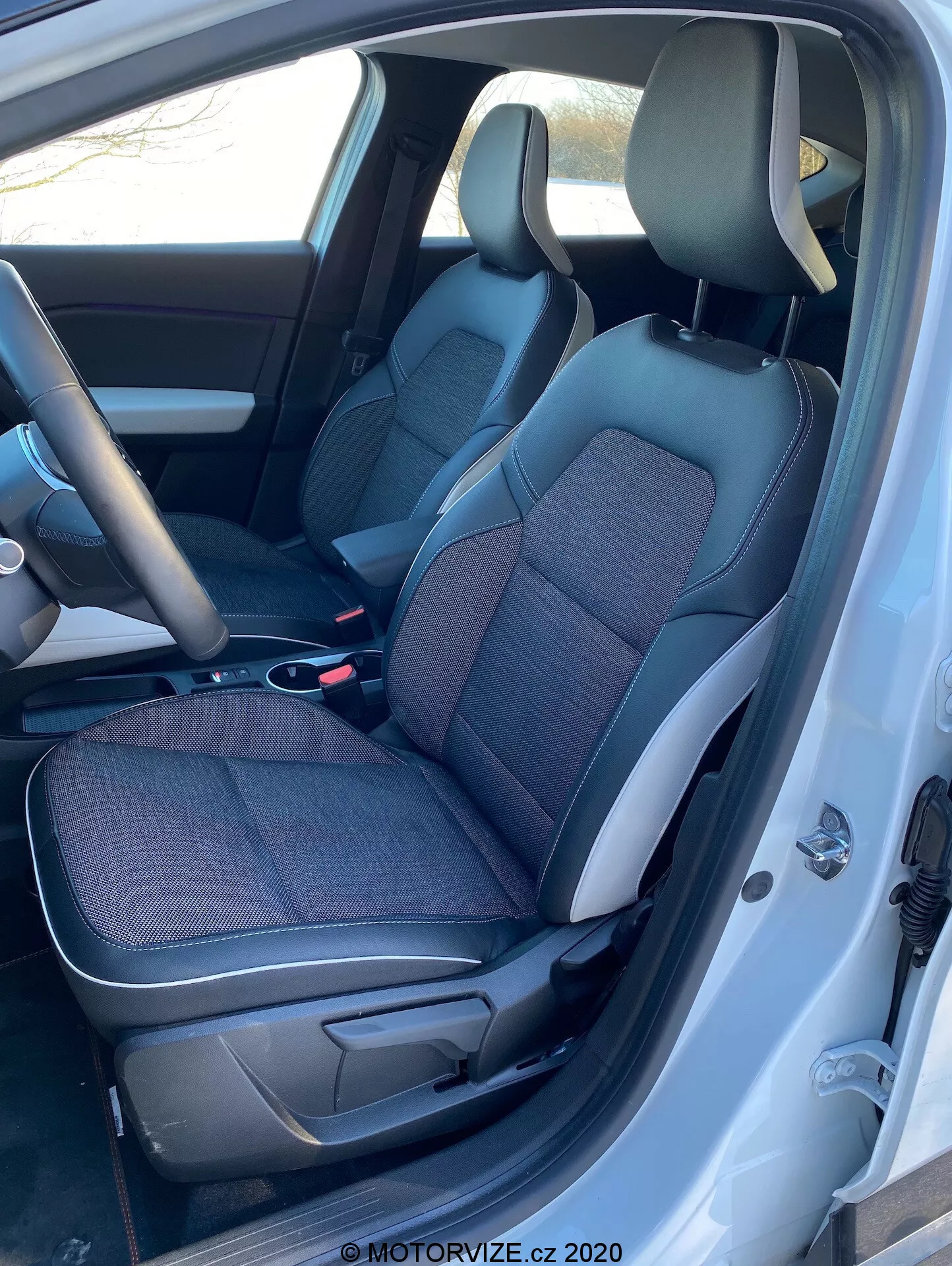 Vista interior del Renault Captur 2020 desde el lado izquierdo con la puerta del conductor abierta, mostrando asientos de tela y cuero en negro y gris, consola central, volante y parte del salpicadero.