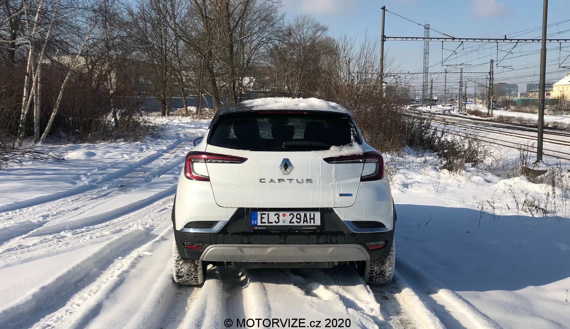 Vista trasera centrada de un Renault Captur 2020 en una carretera nevada. La imagen destaca el diseño trasero distintivo, incluido el esquema de color de dos tonos con carrocería blanca y techo en contraste, iluminación LED trasera moderna, matrícula de estilo europeo, limpiaparabrisas trasero y un solo tubo de escape en el lado derecho. La foto captura líneas eléctricas y árboles en el fondo a la luz del día, lo que sugiere un entorno suburbano o semi-rural.