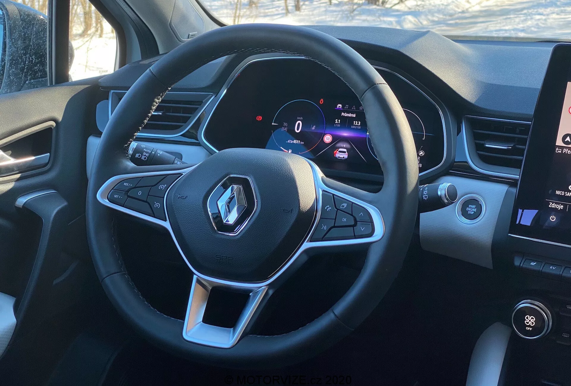 Vista interior del Renault Captur 2020 desde la perspectiva del conductor, destacando el volante multifuncional deportivo de base plana con el logo de Renault y botones de control para funciones de audio y control de crucero. El cuadro de instrumentos digital muestra una velocidad estacionaria de 0 km/h junto a un tacómetro, indicando tecnología moderna e información personalizable para el conductor. A la derecha se puede ver una sección del sistema de infoentretenimiento de pantalla táctil vertical, que muestra los controles de temperatura y la información de los medios, en condiciones de luz diurna que ofrecen una vista realista desde el asiento del conductor.