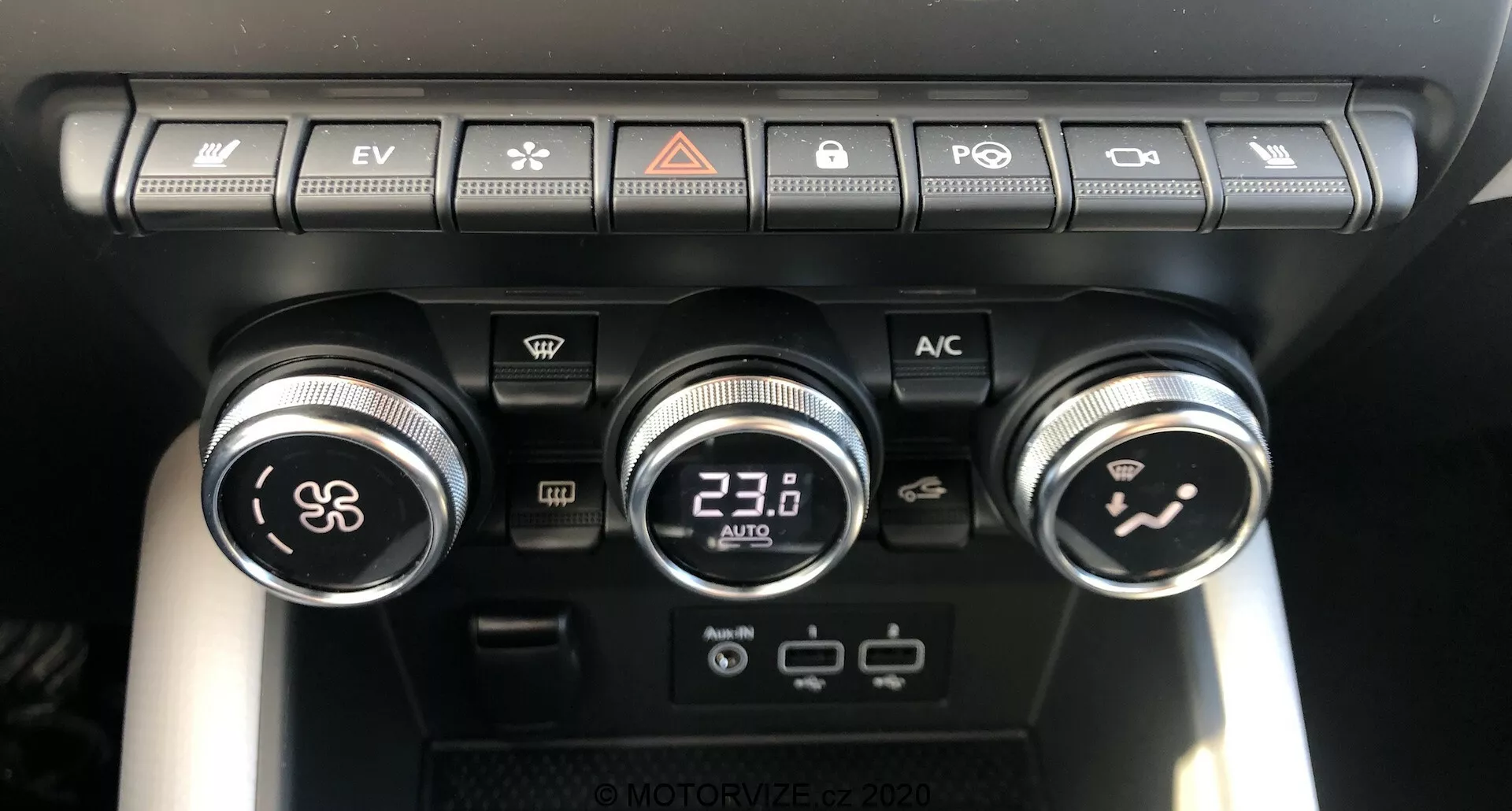 Vista en primer plano del panel de control climático del Renault Captur 2020 en el interior del vehículo, mostrando tres diales y varios botones. El dial izquierdo controla la velocidad del ventilador, el dial central con pantalla digital está ajustado a 23.0°C para la temperatura, y el dial derecho dirige el flujo de aire en la cabina. Sobre los diales, una fila de botones incluye recirculación de aire, desempañado del parabrisas, desempañado de la ventana trasera, sincronización de temperatura, botón de modo automático y botón EV indicando capacidades de vehículo eléctrico. Debajo del dial central hay dos puertos USB y una toma de corriente de 12V, destacando la conectividad moderna y las opciones de carga.