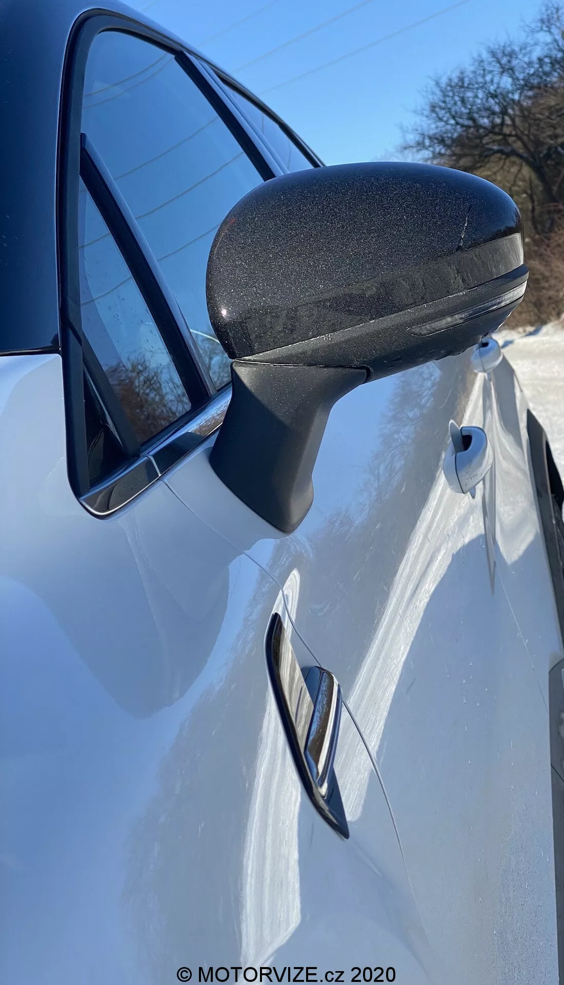 Vista detallada en primer plano del espejo lateral, la puerta y el panel delantero del Renault Captur 2020 blanco. La foto, tomada desde el lado justo delante del espejo lateral en un ángulo ascendente, resalta el diseño moderno y aerodinámico del espejo lateral con una sección inferior texturizada negra para mayor durabilidad. El soporte estilizado se integra perfectamente en el marco de la puerta. La manija de la puerta empotrada y la línea de carácter que la atraviesa sugieren un diseño elegante y deportivo. Un vistazo del faro y un reflejo del cielo claro en la ventana realzan esta presentación dinámica.