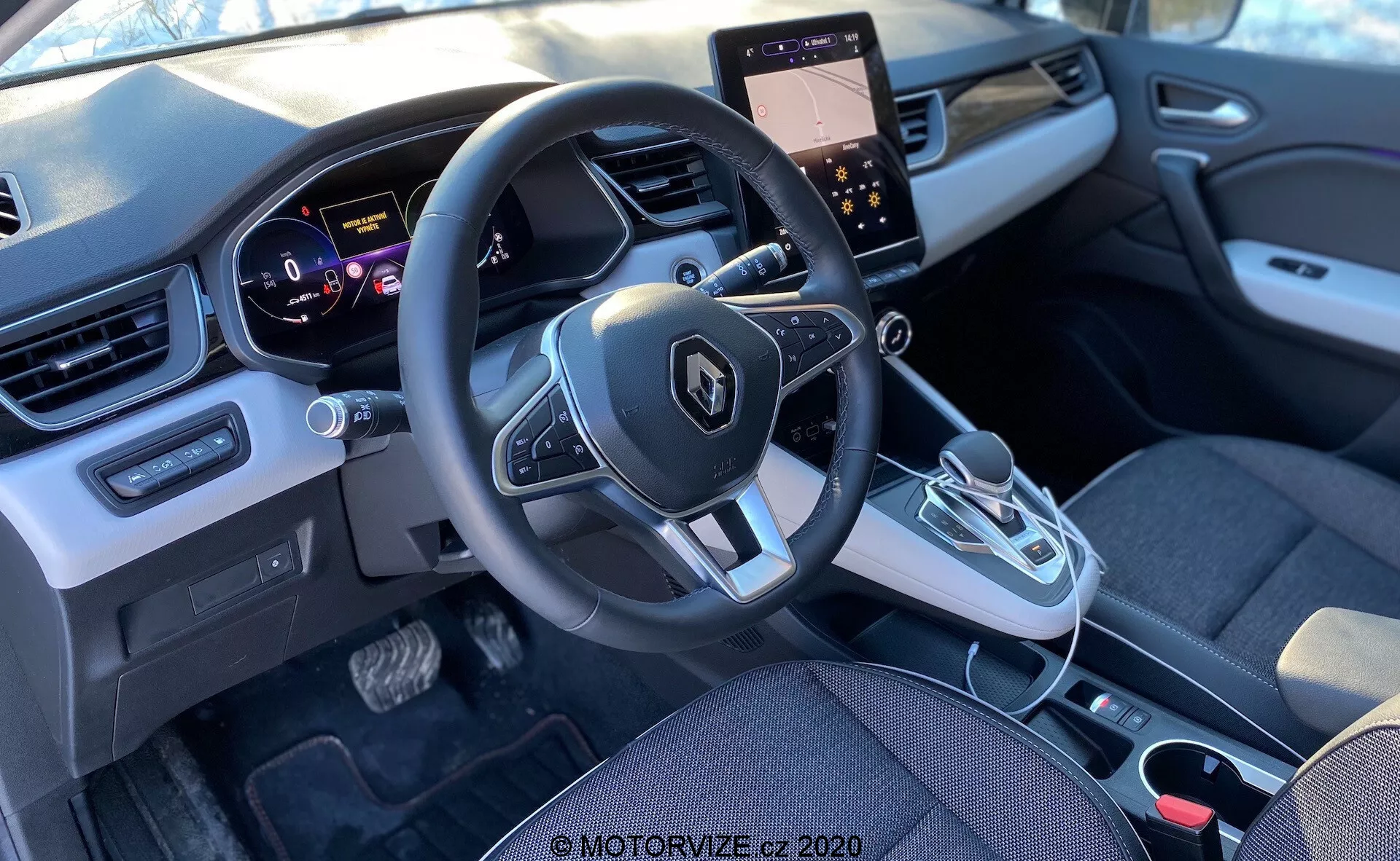 Vista interior de un Renault Captur 2020 desde el asiento trasero, enfocándose en el área de la cabina frontal, incluyendo el tablero, volante con controles integrados, cuadro de instrumentos, consola central con pantalla táctil y controles de clima, palanca de cambios automática y diseño moderno con elementos de tela y plástico.
