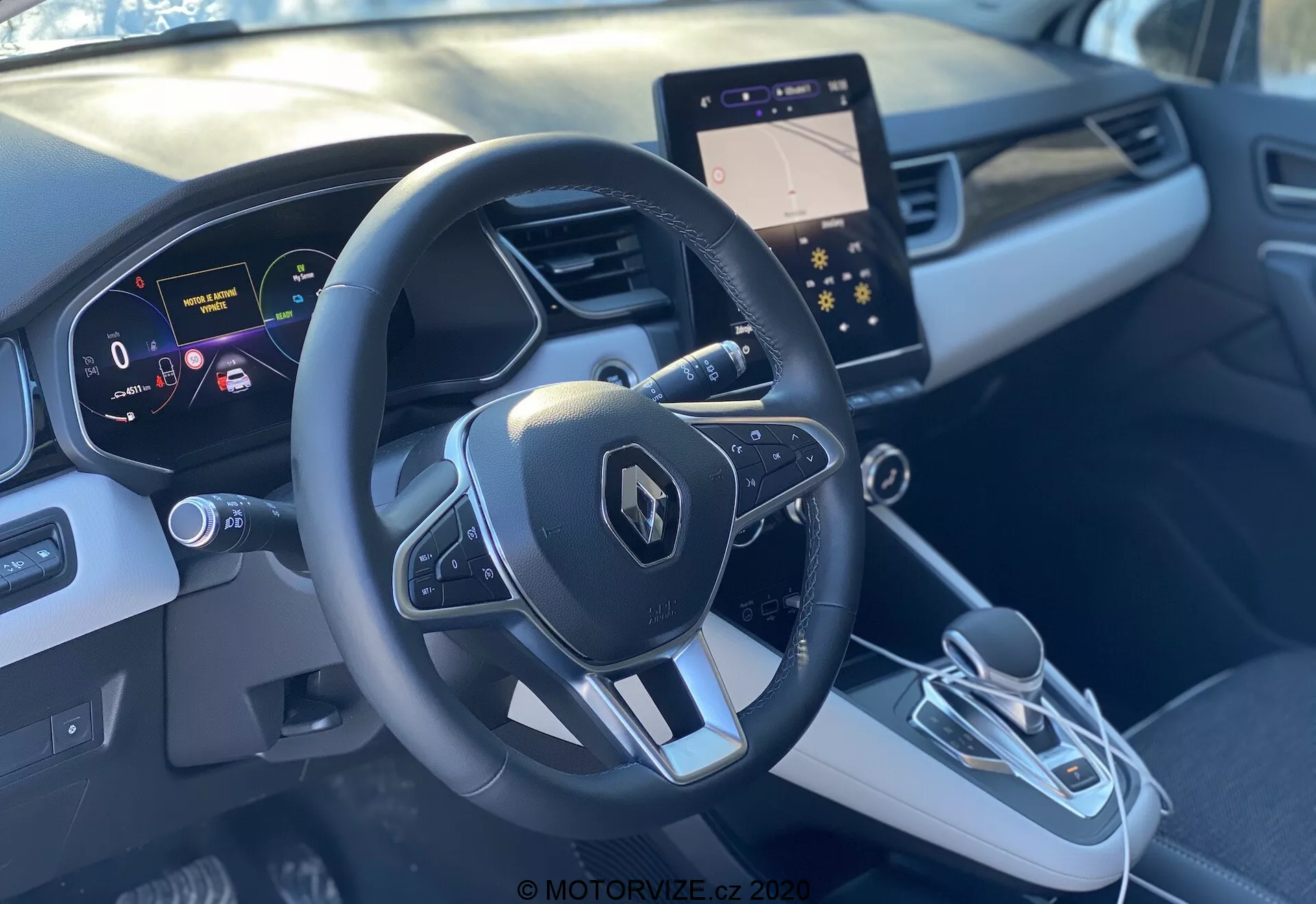 Vista interior del Renault Captur 2020 desde el asiento del acompañante delantero, mostrando el volante deportivo de tres radios con el emblema de Renault y botones multifuncionales, panel de instrumentos digital con pantalla de alta resolución mostrando la velocidad y ayudas de navegación, y una elegante consola central. La consola central cuenta con una moderna pantalla de infoentretenimiento, palanca de cambio electrónica y botón de freno de estacionamiento electrónico. El interior bicolor en gris con diseño contorneado y adornos proporciona una sensación premium.
