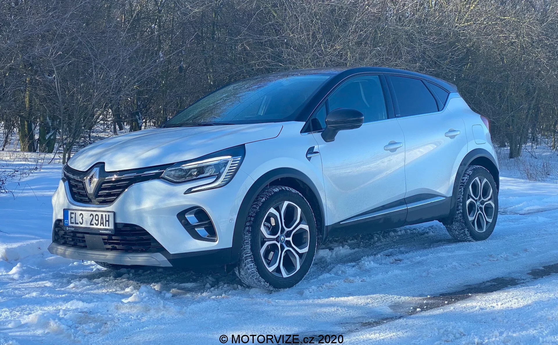 Vista desde un ángulo bajo de un SUV Renault Captur 2020 sobre una superficie nevada, destacando su perfil de cuarto delantero. La luz brillante del día y el cielo despejado resaltan los elementos de diseño moderno como la prominente parrilla frontal con insignia, las luces diurnas LED, las intrincadas ruedas de aleación de dos tonos, la carrocería esculpida con arcos de rueda pronunciados, los espejos del color de la carrocería con señales de giro integradas, las ventanas tintadas para mayor privacidad y las manijas de las puertas al ras. La foto enfatiza la construcción premium del vehículo y sus posibles capacidades de conducción en invierno.