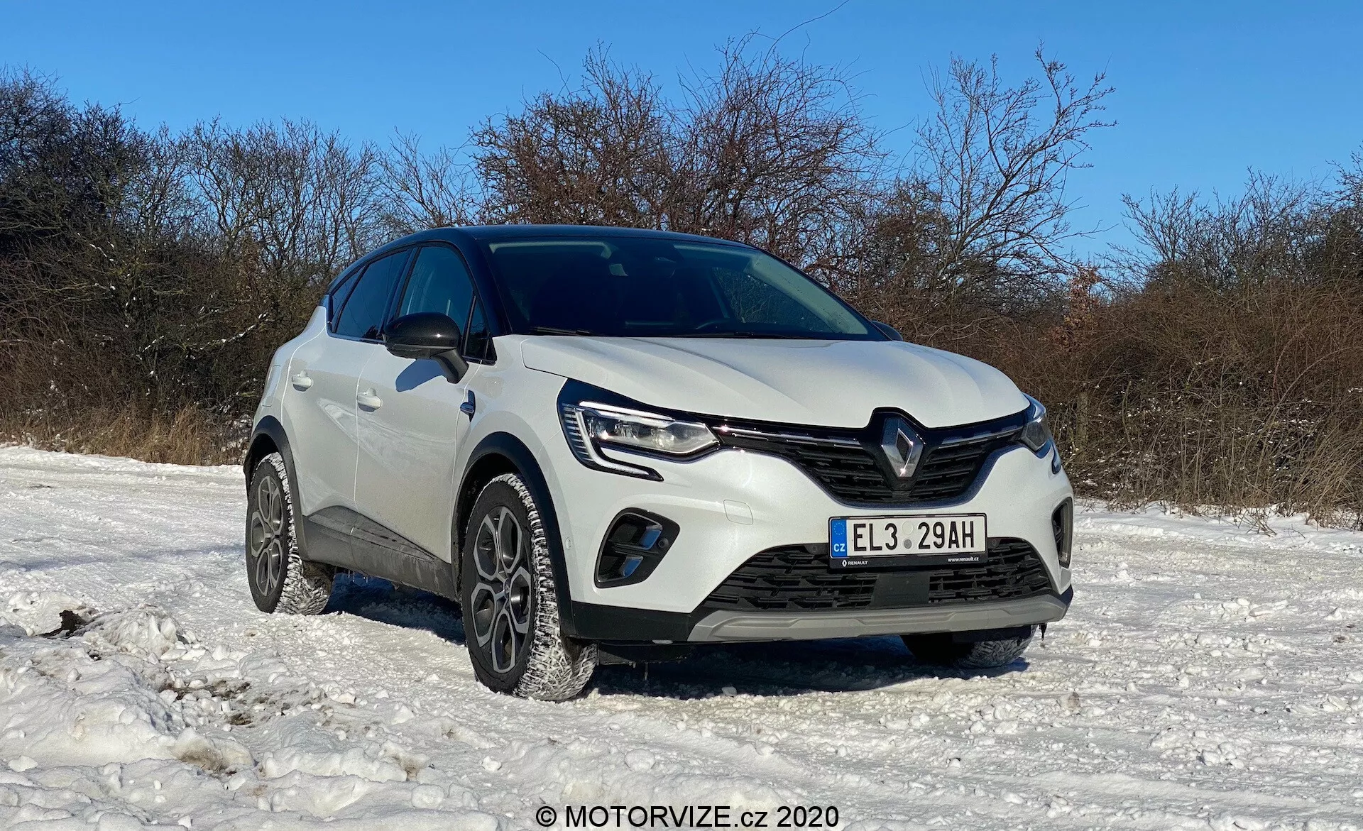 Renault Captur 2020 blanco estacionado en un terreno cubierto de nieve bajo un cielo despejado, mostrado desde un ángulo de tres cuartos delanteros bajo, mostrando la parrilla delantera, faros con luces diurnas LED, luces antiniebla, parachoques delantero, neumáticos de invierno, logo distintivo de Renault, líneas esculpidas en el capó, altura elevada, posibles sensores de asistencia de estacionamiento, espejos laterales y estado general limpio y bien mantenido, con la marca de agua MOTORVIZE.cz © 2020.