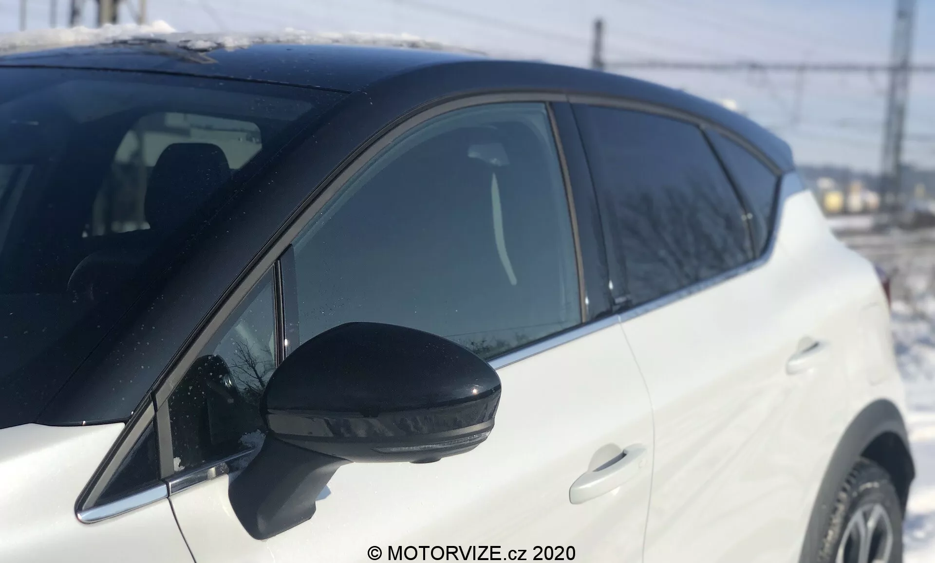 Vista lateral en primer plano de un Renault Captur 2020 blanco, enfocándose desde cerca del espejo lateral hasta antes de la rueda delantera. Espejo lateral negro prominente con indicador de señal de giro integrado, tomado a la luz del día con luz natural. Ángulo ligeramente bajo que enfatiza la sección media, poca profundidad de campo. Fondo desenfocado con elementos discernibles como una cerca, líneas eléctricas y una estructura que indican un entorno exterior. Pintura blanca brillante del vehículo y manijas de las puertas a juego con el color, mostrando un diseño moderno bien mantenido. Línea de ventana ascendente hacia la parte trasera, indicando un estilo dinámico.