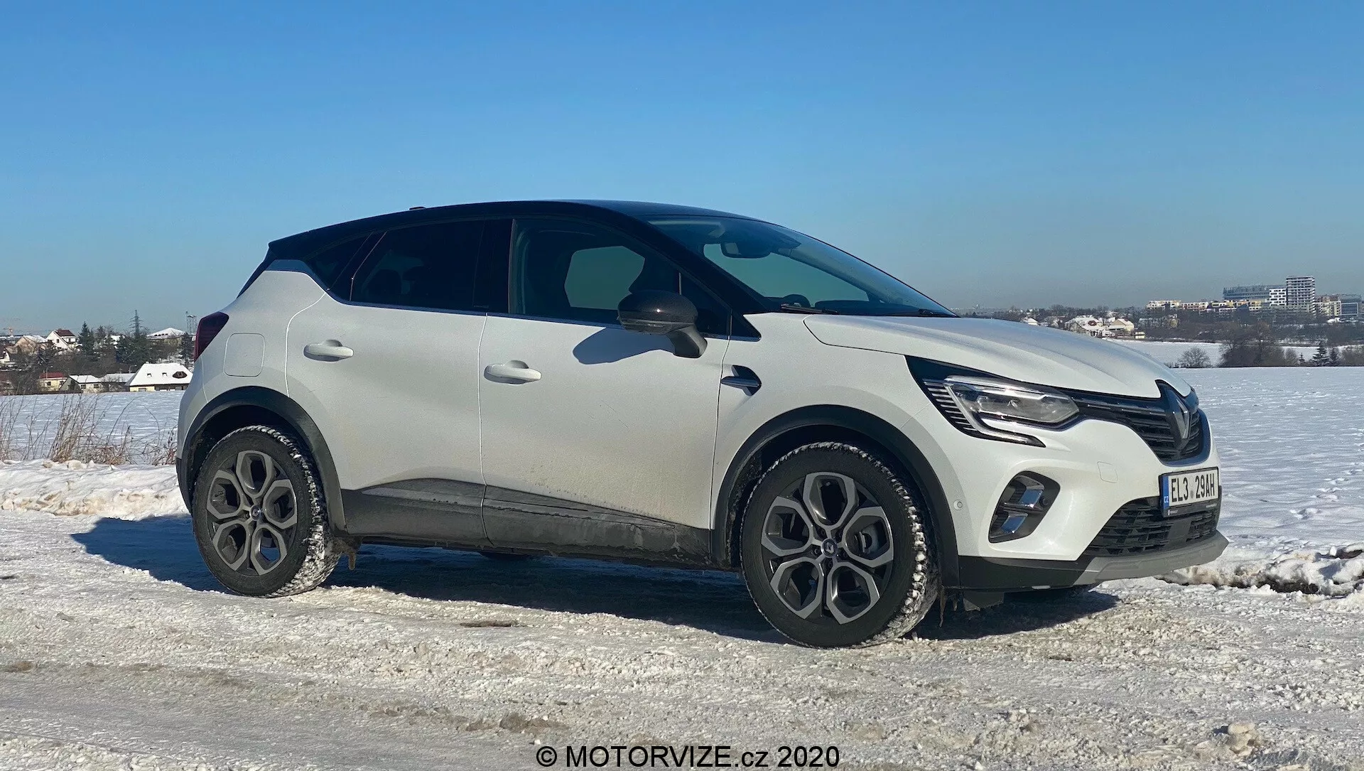 Renault Captur 2020 moderno SUV crossover posicionado en un terreno helado y nevado con un cielo claro de fondo. El lado del pasajero mira a la cámara en un ángulo leve, mostrando el frente, el lado y parcialmente la parte superior del vehículo. Cuenta con luces diurnas LED, parrilla frontal integrada con el emblema del fabricante, pintura bicolor clara y negra, llantas de aleación de cinco radios, molduras protectoras de plástico, manijas de puertas elegantes y empotradas y una línea de ventana afilada con un alerón trasero. Sugiere tecnología avanzada como iluminación LED y entrada sin llave, ambientado en un paisaje de clima frío cerca de una zona urbana.