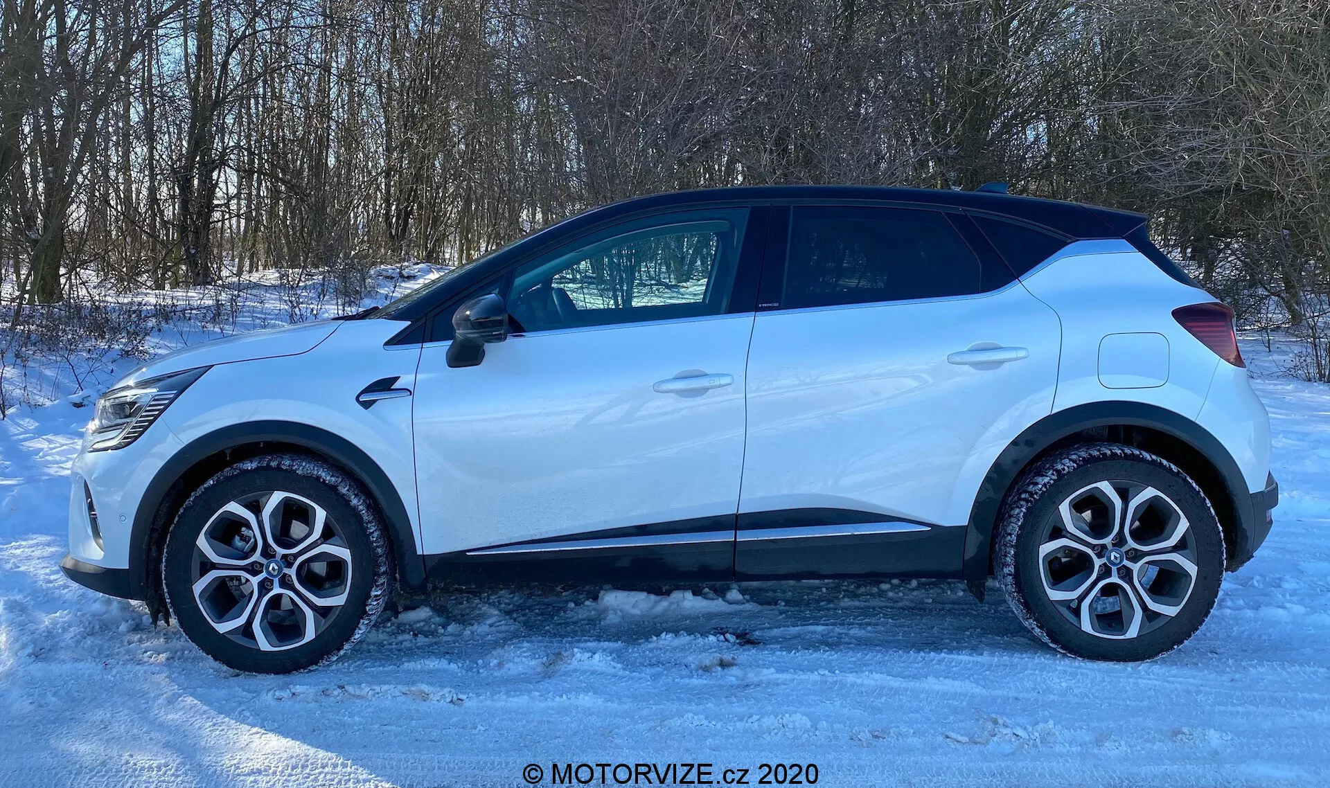 Vista lateral de un Renault Captur 2020 estacionado sobre una superficie helada o nevada. El perfil del coche muestra llantas de aleación, neumáticos de carretera estándar, cuatro puertas, ventanas laterales, incluyendo una pequeña ventana trasera, y un espejo retrovisor lateral con un indicador de señal de giro integrado. La línea del techo está ligeramente inclinada hacia la parte trasera, con revestimientos plásticos alrededor de los arcos de las ruedas y la parte inferior de las puertas, lo que sugiere un diseño preparado para todo terreno. La escena bien iluminada, probablemente por luz solar natural, define la forma del coche contra un entorno rural o suburbano con árboles en el fondo. La imagen tiene una marca de agua que dice 'MOTORVIZE.cz 2020'.