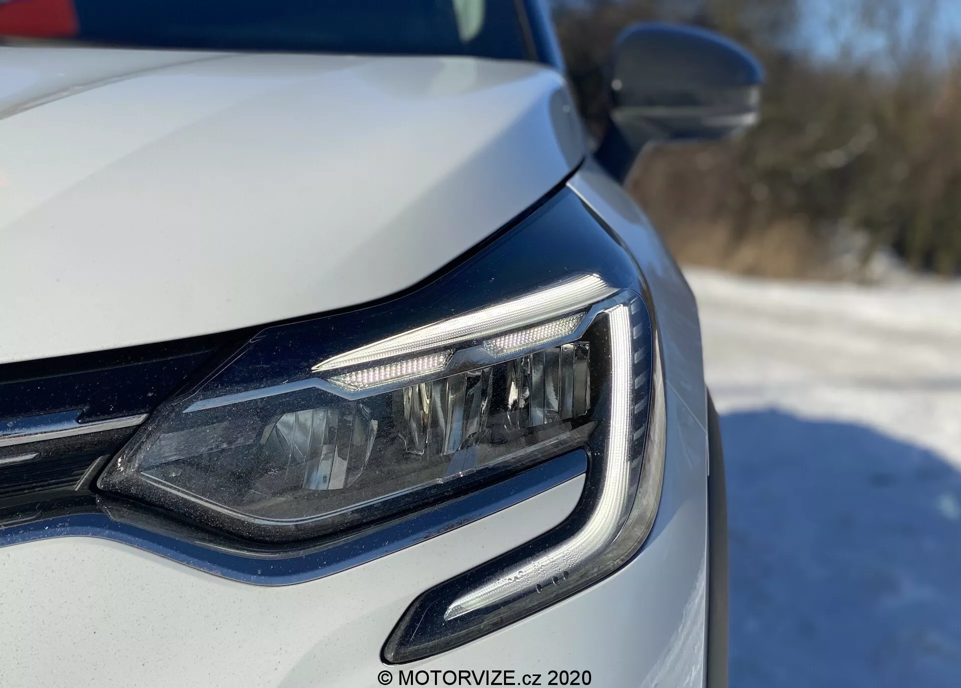 Vista de cerca del rincón delantero izquierdo de un coche moderno, enfocándose en el faro LED y la parrilla, tomada desde un ángulo bajo cerca del nivel del faro. Las luces diurnas (DRL) tienen un diseño curvo que abraza la unidad del faro. Posibles faros adaptativos o luces de esquina visibles, con acabado de pintura metálica y señales de giro integradas en el espejo retrovisor exterior. La iluminación natural brillante sugiere un entorno al aire libre durante el día.