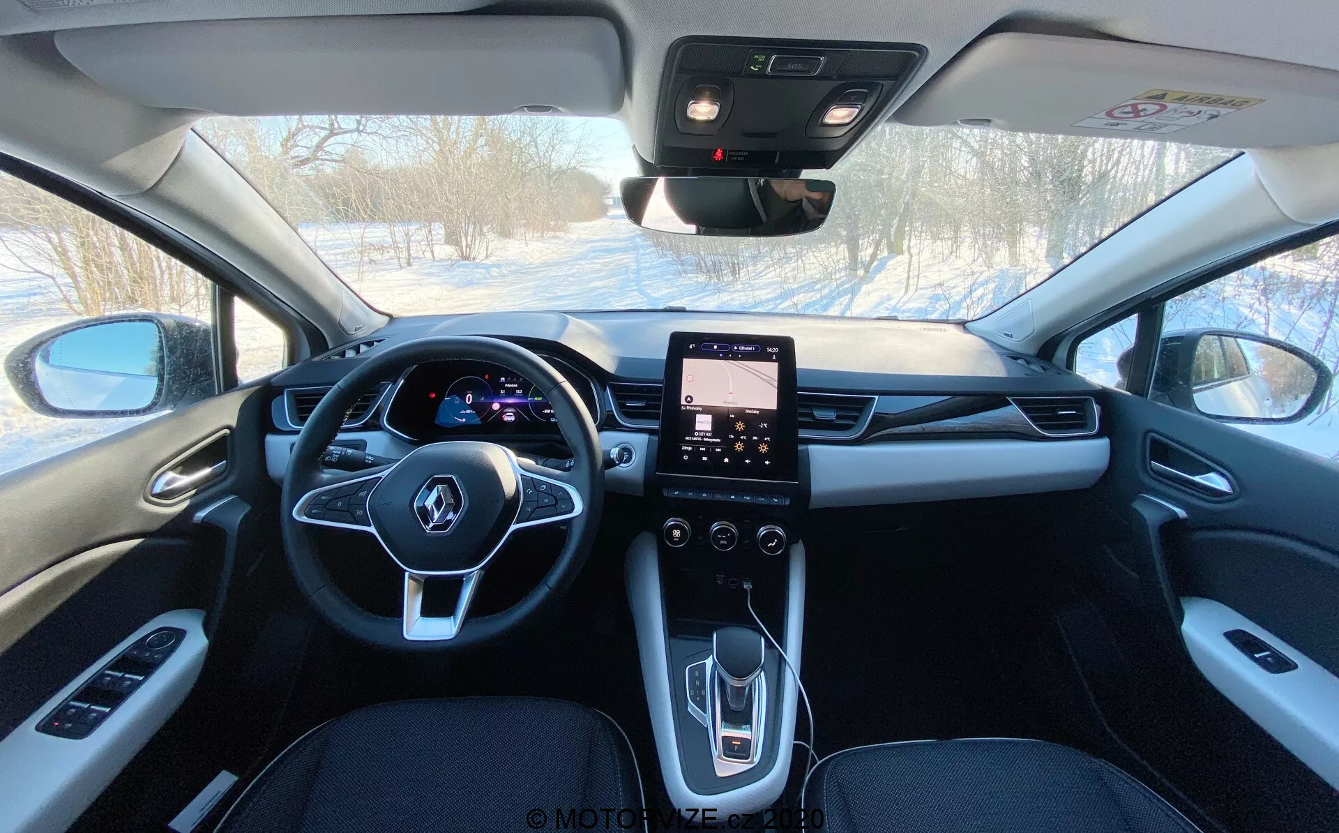 Vista interior de un Renault Captur 2020 desde la zona de asientos traseros mirando hacia adelante, mostrando su tapicería de color oscuro, espejo retrovisor central, panel de instrumentos digital iluminado detrás del volante multifunción, sistema de infoentretenimiento con pantalla táctil en la consola central y controles para ventanas eléctricas, espejos laterales, control de clima, y probablemente para el techo solar y botón SOS de emergencia.