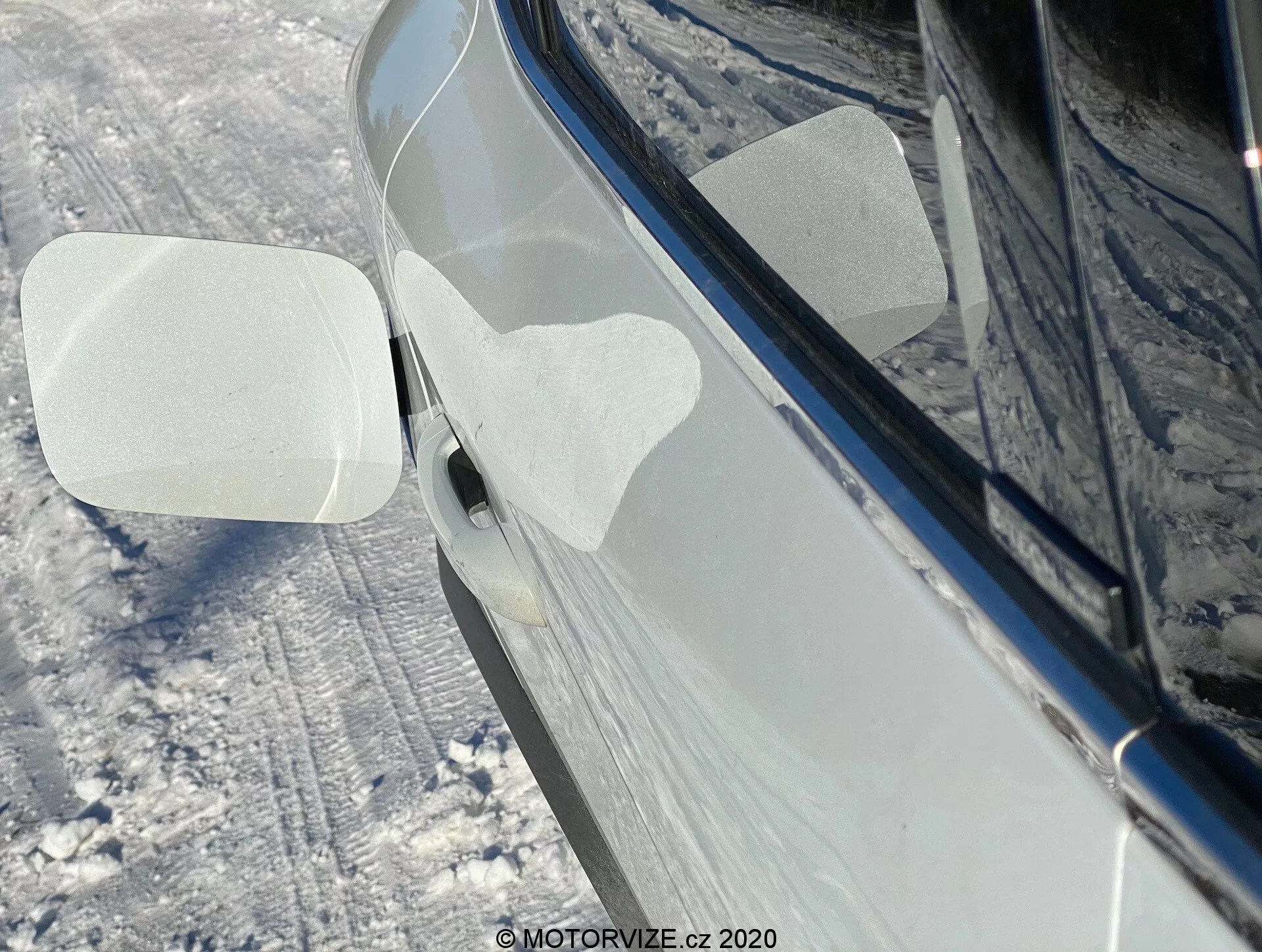 Vista lateral en primer plano de un Renault Captur 2020 en un entorno nevado al aire libre. El enfoque está en el espejo retrovisor derecho, que está doblado hacia el frente, posiblemente indicando una característica de plegado automático o manual. La carcasa del espejo tiene dos láminas protectoras transparentes adheridas en forma de paralelogramos, probablemente para protección durante la entrega o el lavado. El coche, de un tono oscuro brillante como negro o azul oscuro, parece bien mantenido con reflejos que sugieren una superficie limpia y pulida. El suelo alrededor y los lados del coche están cubiertos de nieve. También es visible parte de la manija de la puerta delantera, con un botón de entrada sin llave o sensor táctil.
