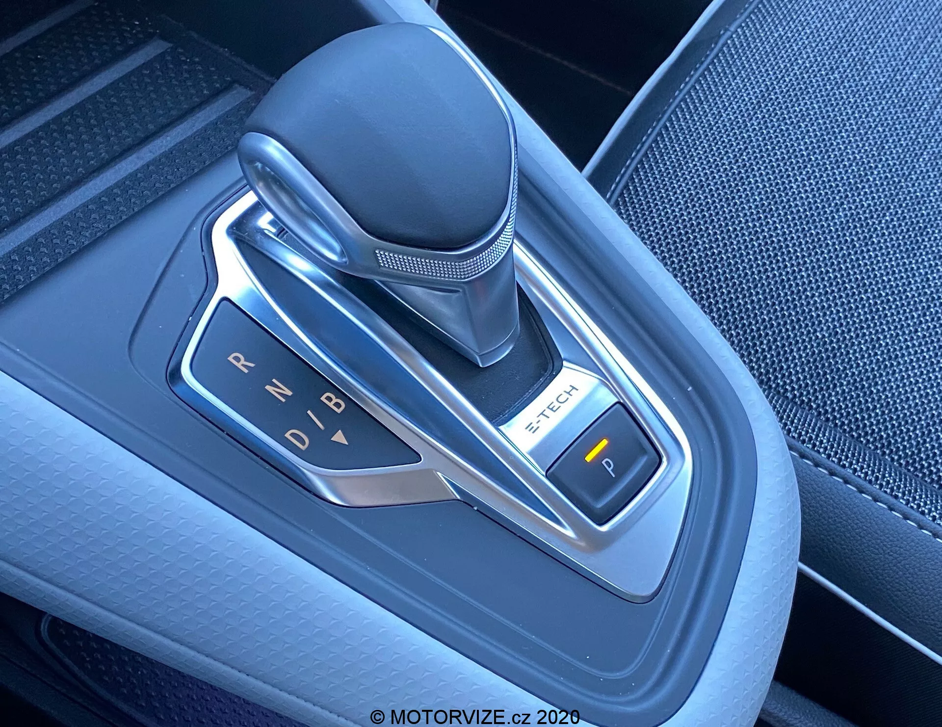 Imagen en primer plano de una moderna palanca de cambios automática en un Renault Captur 2020, ubicada en la consola central. El selector de marchas electrónico está posicionado verticalmente y hecho de cuero, plástico y detalles metálicos. La fotografía, tomada desde un ángulo superior ligeramente desde la derecha, resalta las indicaciones de cambio (P, R, N, D, B) iluminadas con luces LED. A la derecha es visible un botón etiquetado con 'P', probablemente para el freno de mano electrónico. El área alrededor de la palanca de cambios presenta plástico texturizado de alta calidad y detalles metálicos, reflejando la avanzada tecnología y el diseño contemporáneo del interior del vehículo.