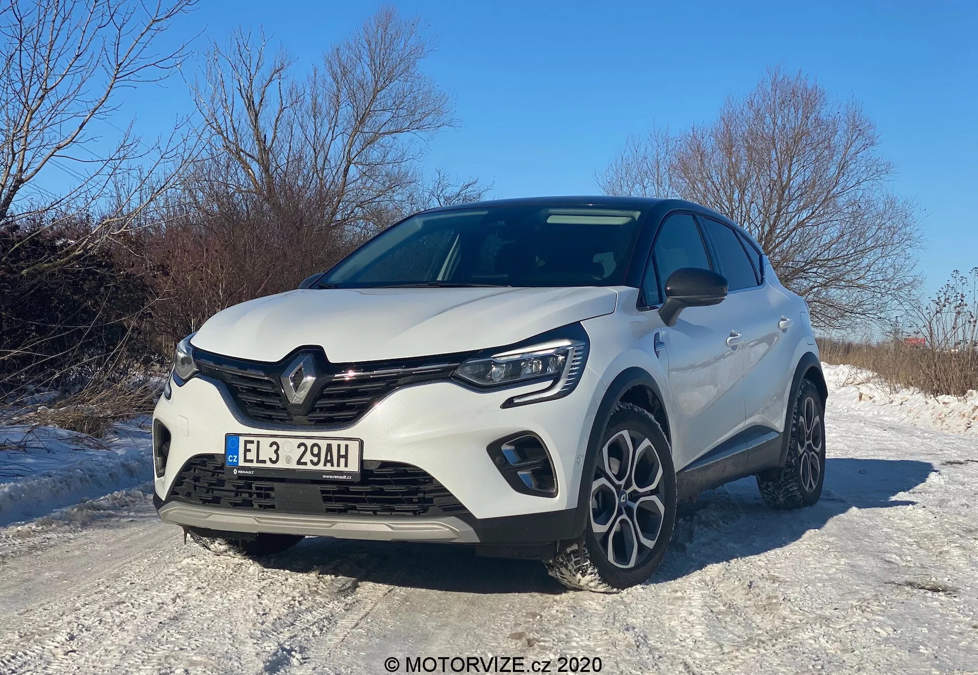 Vehículo hatchback moderno Renault Captur 2020 capturado desde un ángulo bajo de tres cuartos frontal, mostrando la parrilla frontal, el capó, los faros LED, el parachoques delantero con luces antiniebla integradas y un perfil lateral parcial. Los elementos de diseño incluyen una parrilla prominente con el emblema del fabricante, líneas esculpidas en el capó y llantas de aleación de múltiples radios, todo bajo un cielo claro de día, reflejando una estética deportiva y aerodinámica, posiblemente destacando un nivel de equipamiento mejorado o un paquete deportivo. El automóvil está situado en una carretera nevada, lo que sugiere características adecuadas para la conducción en invierno.