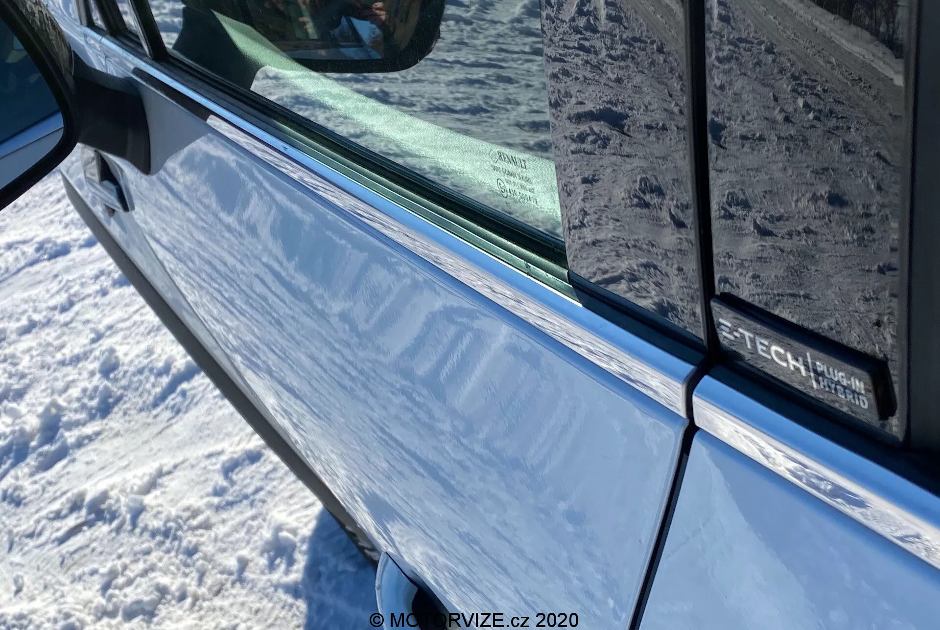 Primer plano del Renault Captur 2020 en azul metálico, centrado en la puerta del conductor y el espejo lateral con señal de giro integrada. La imagen destaca el área de la puerta con un borde negro a lo largo del borde de la ventana tintada, que refleja el entorno circundante. En la puerta se puede ver parcialmente un distintivo que indica un posible paquete de tecnología o acabado.
