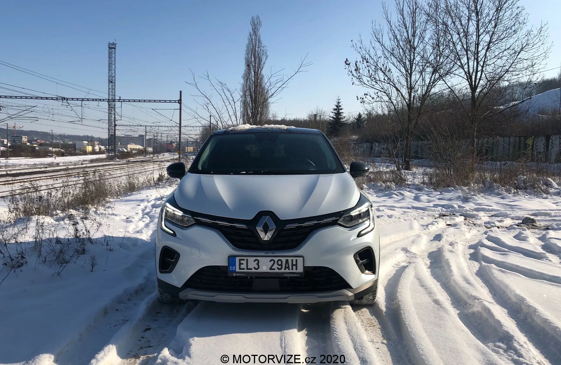 Vista frontal directa del Renault Captur 2020 en una carretera cubierta de nieve, mostrando en detalle la parrilla delantera, los faros, el capó esculpido y el parachoques delantero a la luz del día. Las huellas de los neumáticos son visibles, evidenciando movimiento reciente. El diseño del vehículo sugiere una aerodinámica estilizada, con un emblema de marca prominente y un sistema de iluminación avanzado. Podrían estar integrados sensores o cámaras de asistencia al conductor en el parachoques delantero.