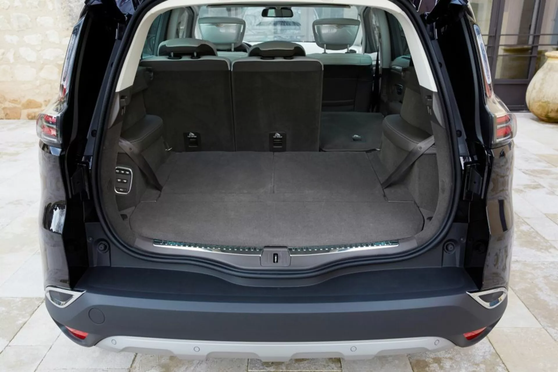Vista interior trasera del Renault Espace 2015 mostrando un área de carga limpia con el maletero abierto, que cuenta con un piso de carga plano, asientos traseros abatibles 60/40, una toma de corriente, ganchos para carga, botón de control para el maletero electrónico y una franja de adorno cromada, bajo iluminación natural.