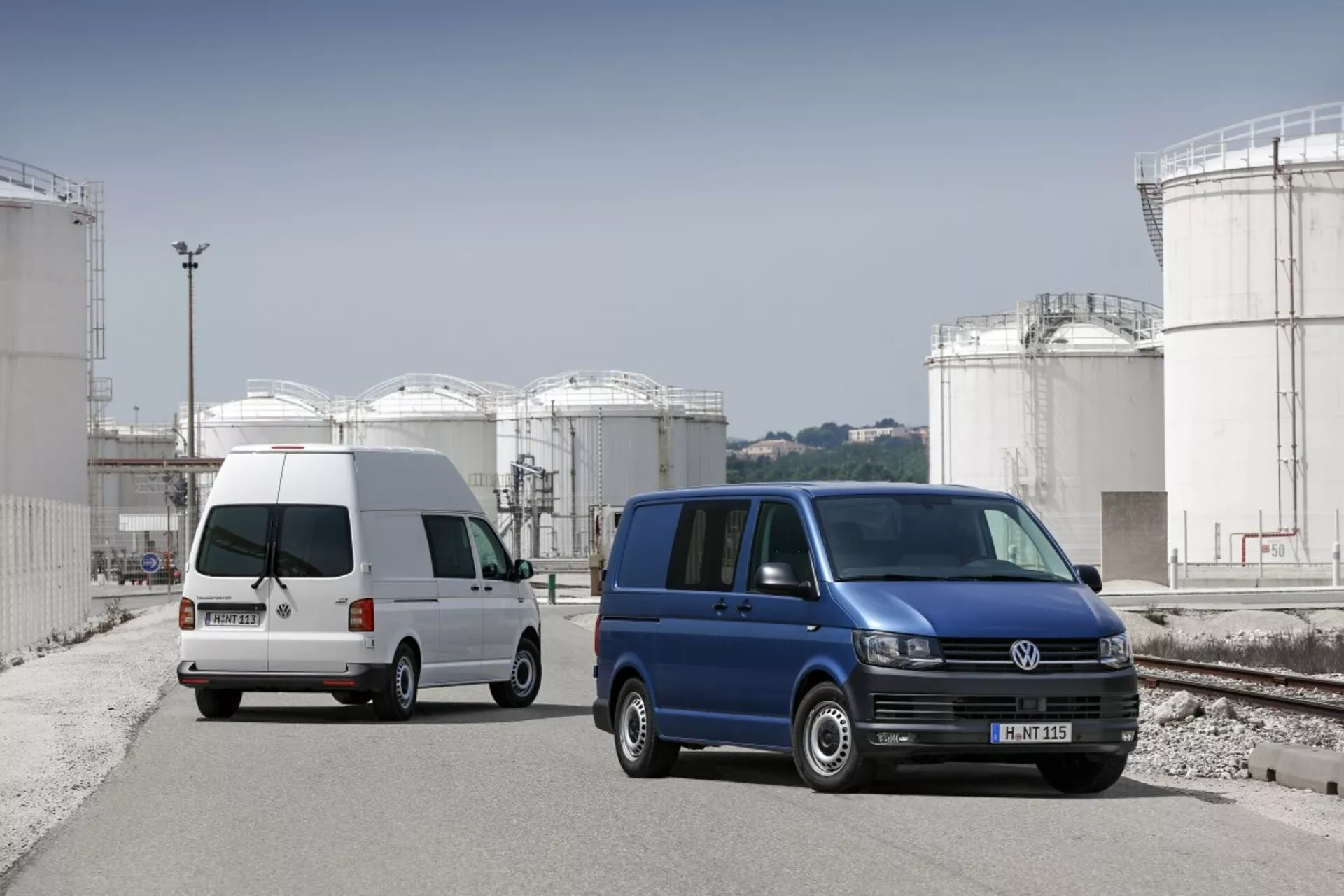 Dos furgonetas Volkswagen Transporter aparcadas en un sitio industrial con grandes tanques de almacenamiento en el fondo, mostrando su diseño de utilidad comercial, puertas laterales correderas y ruedas de aleación estándar.