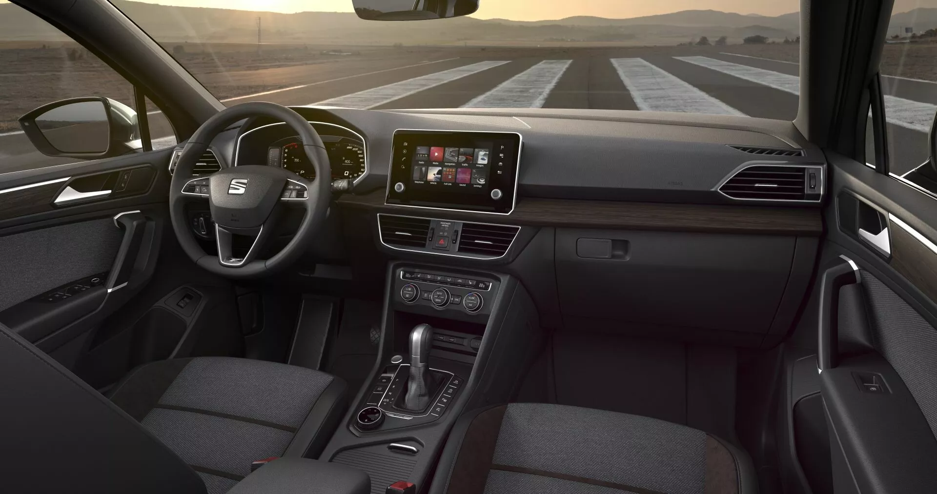 Perspectiva interior del Seat Tarraco 2019 desde el asiento trasero, mostrando el volante multifunción, pantalla táctil central de infotenimiento, botones de control climático, palanca de cambio automático y asiento del pasajero tapizado en tela, bajo una iluminación interior sutil