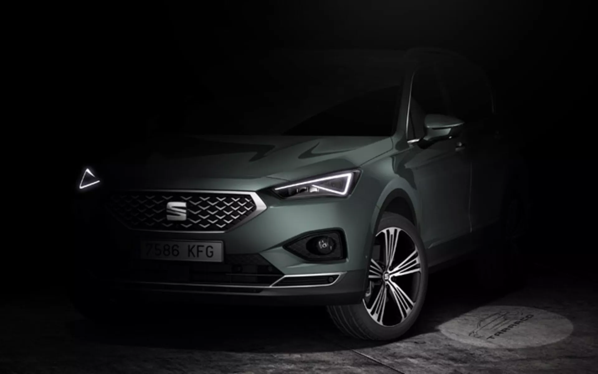 Seat Tarraco 2019 mostrado en un ambiente oscuro y tenue que enfatiza su elegante frente, parrilla geométrica, faros LED con distintivas luces diurnas y capó esculpido, junto a una vista detallada de la llanta de aleación, resaltando los atributos de lujo y tecnología moderna del coche