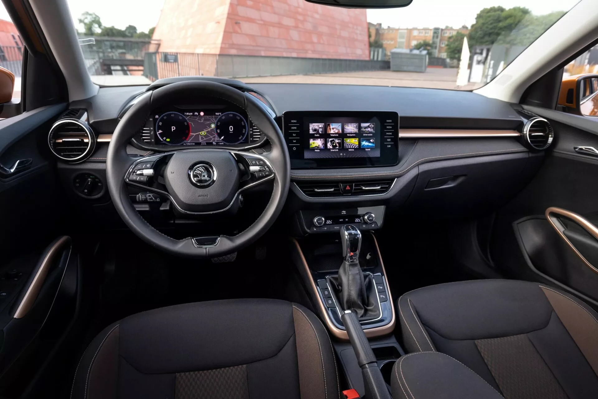 Vista interior del Skoda Fabia 2021 desde el asiento trasero, que muestra un volante de tres radios con controles, un cuadro de instrumentos digital que muestra el velocímetro y el tacómetro, y un sistema de infoentretenimiento central con pantalla táctil con iconos de navegación y música. Rejillas de ventilación circulares y horizontales con acabado que imita madera o metal, transmisión automática con freno de mano electrónico en la consola central, asientos delanteros deportivos con tapicería de cuero y costuras de contraste. Ambiente moderno y amplio espacio interior que indican lujo y deportividad.