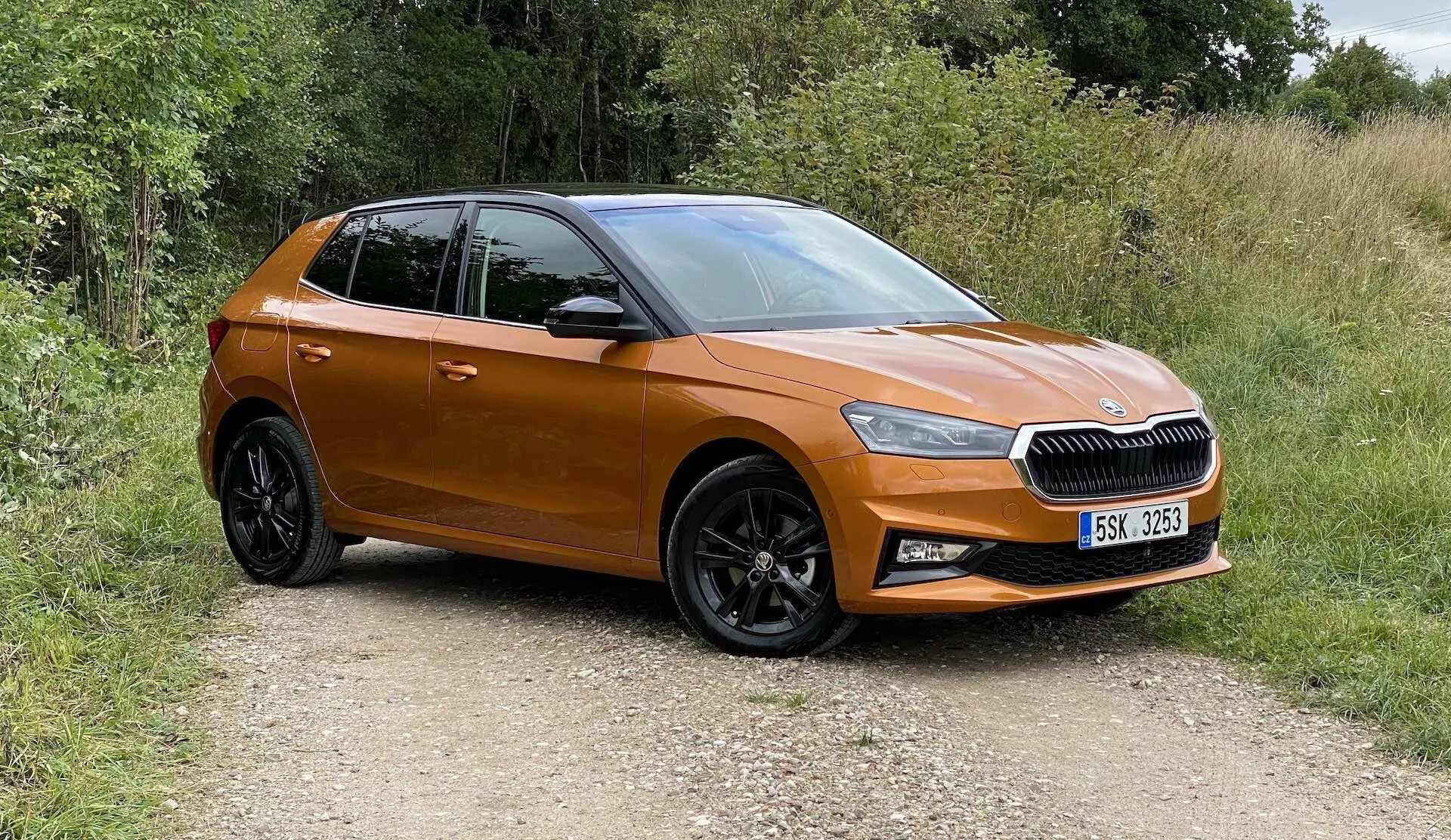 Imagen de un moderno hatchback Škoda Fabia 2021, capturada desde una vista frontal a tres cuartos, que muestra el lado izquierdo delantero, la puerta del conductor, el capó izquierdo y parte del techo. El vehículo está en un camino no pavimentado rodeado de vegetación, indicando un entorno rural. Las características visibles incluyen una parrilla horizontal con emblema central, faros LED afilados, pintura metálica naranja, espejos laterales del color de la carrocería, ruedas de aleación de diseño multirradio negro con neumáticos con perfil y puertas contorneadas con líneas aerodinámicas. El coche parece nuevo, compacto y sin modificaciones.