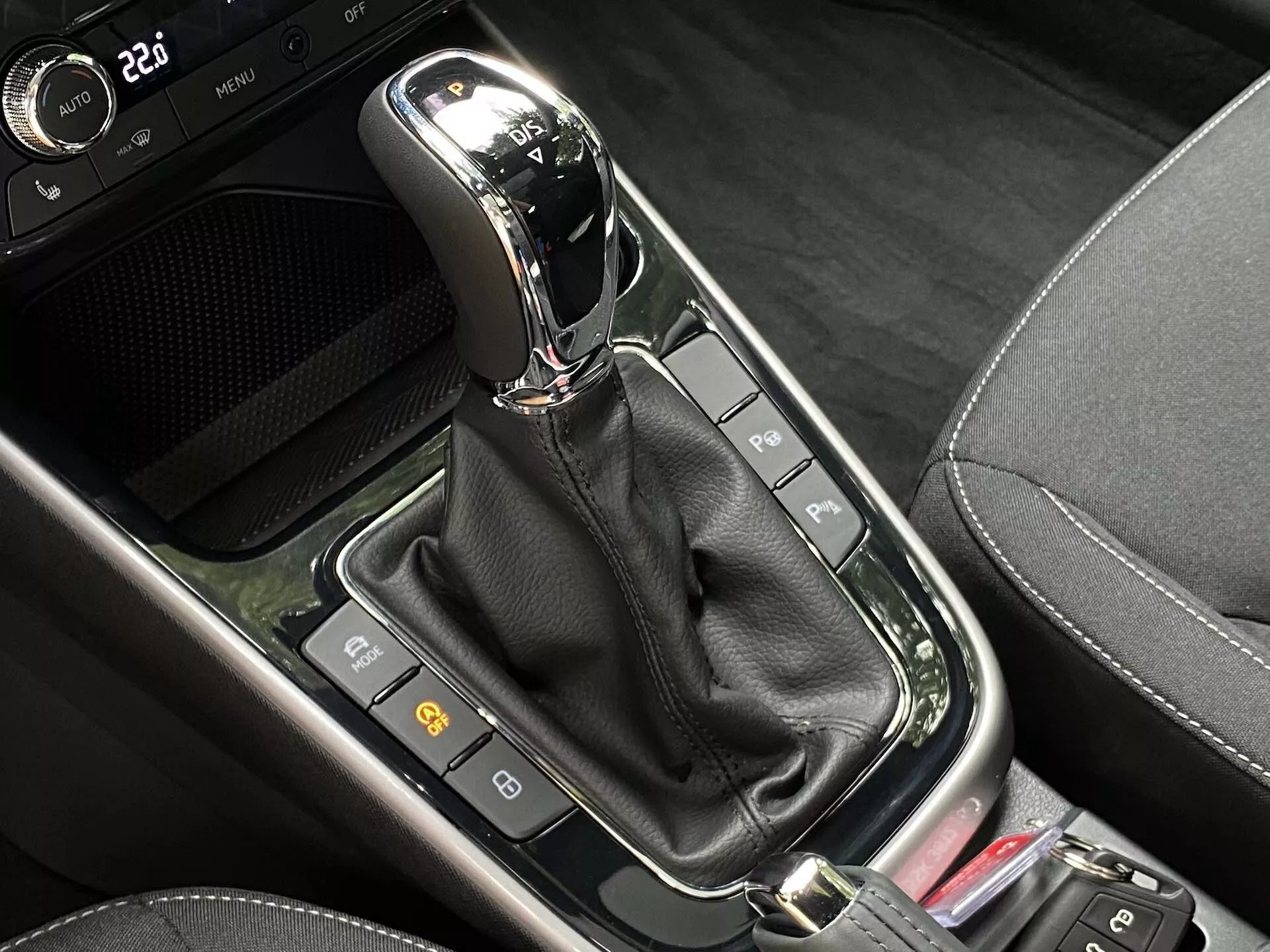Vista de cerca del selector de cambios automático en la consola central del Skoda Fabia 2021. El selector moderno, ubicado verticalmente en la posición 'P' (aparcar), cuenta con un acabado cromado o metálico en la parte superior y está encapsulado en un panel negro brillante. Debajo, una funda de cuero negro oculta el mecanismo. Alrededor del selector, hay una serie de botones e indicadores, incluidos dos a la izquierda que probablemente están relacionados con características de seguridad del coche, y parte de un botón rojo a la derecha. La zona combina diversos materiales negros, incluyendo superficies brillantes y patrones que imitan la fibra de carbono, sugiriendo un diseño contemporáneo de alta tecnología.
