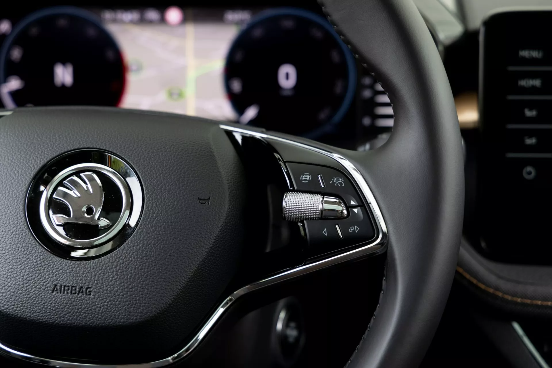 Vista en primer plano del interior del Skoda Fabia 2021 desde la perspectiva del conductor, enfocando en los controles del volante del lado derecho. El volante revestido en cuero presenta un emblema plateado central, con el logo de Skoda y la palabra 'AIRBAG' grabada debajo. El grupo de controles del lado derecho incluye botones para el sistema de entretenimiento, llamadas telefónicas manos libres o comandos de voz, con palancas de cambio visibles detrás del volante. El fondo desenfocado muestra el panel de instrumentos con el tacómetro y el velocímetro, y una pantalla digital de asistencia al conductor. Consola central o sistema multimedia parcialmente visible a la derecha.