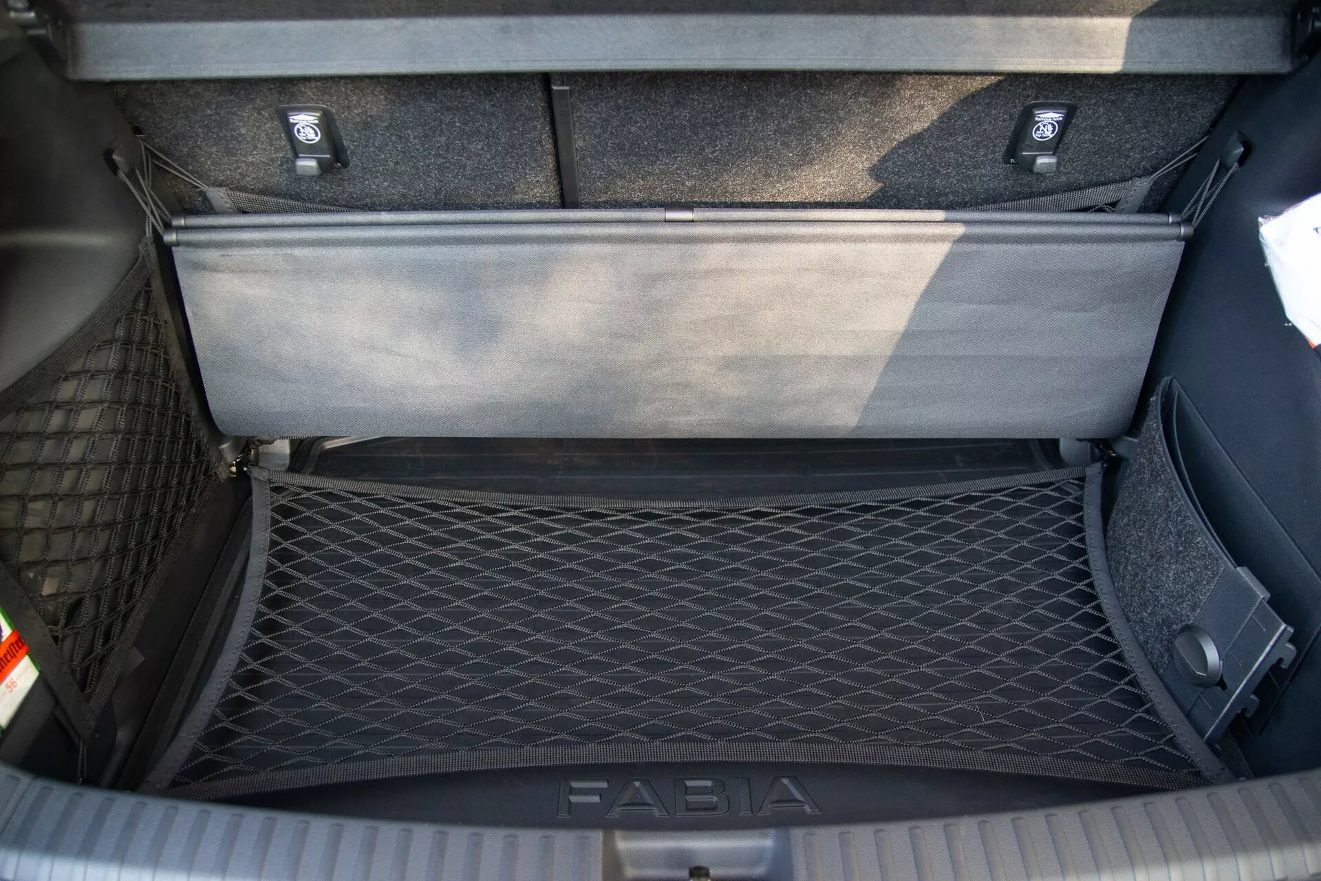 Vista desde arriba del área del maletero de un Skoda Fabia 2021, mostrando el compartimento de almacenamiento interior detallado con una red de carga en el lado izquierdo para la sujeción de objetos y un panel de suelo flexible parcialmente levantado que revela un área de almacenamiento adicional debajo. El maletero cuenta con alfombras duraderas y una placa de umbral con la marca 'FABIA' para protección durante la carga y descarga.