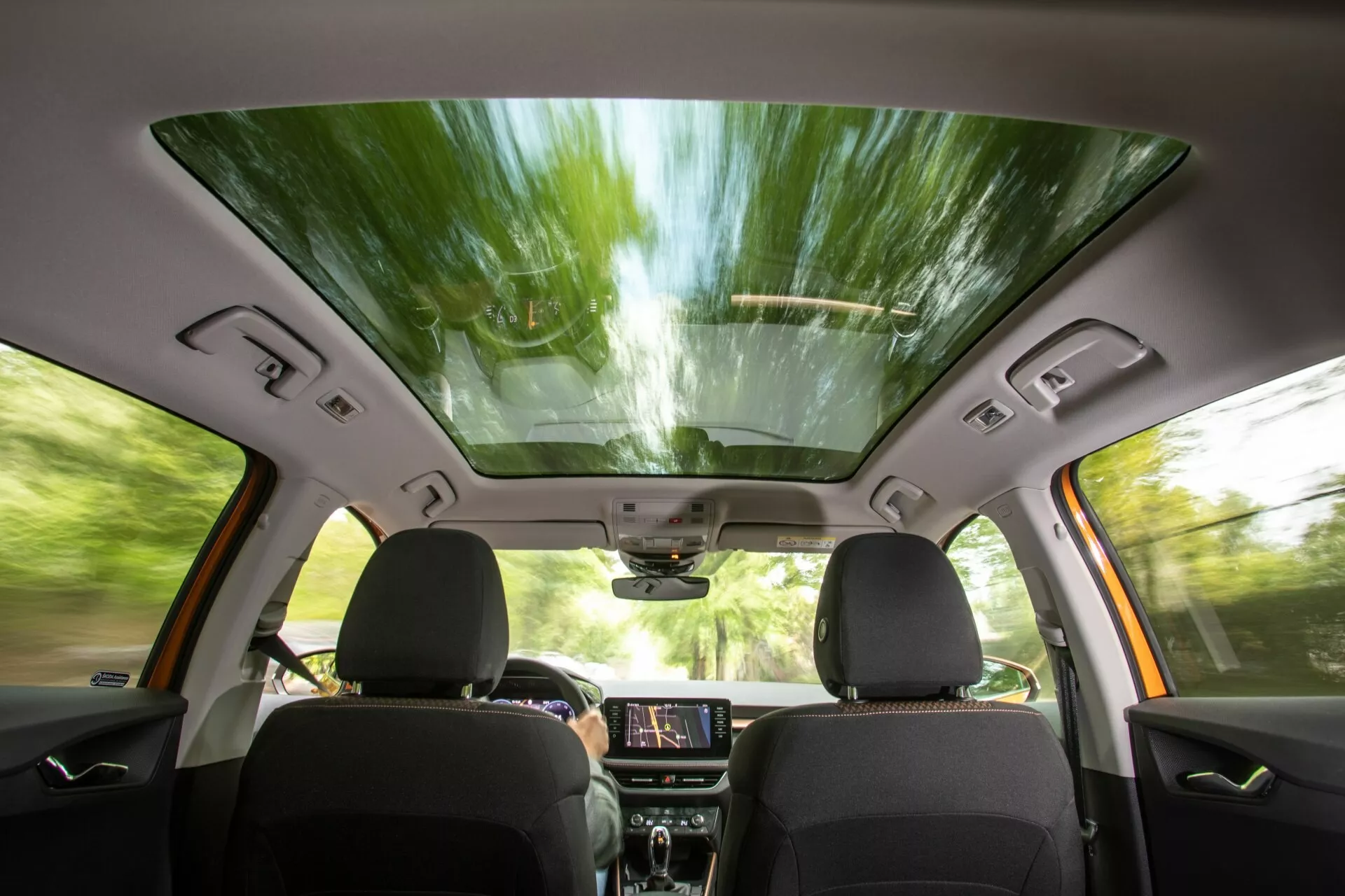 Vista interior del Skoda Fabia 2021 desde la perspectiva del asiento trasero, mostrando los asientos delanteros con tapicería oscura, techo solar panorámico, pantalla táctil de infoentretenimiento iluminada en el tablero, paneles de control, altavoces integrados en las puertas y controles de ventanas, espejo retrovisor con posibles funciones de alta tecnología y una consola superior para controles adicionales, lo que sugiere una experiencia de conducción moderna, cómoda y mejorada tecnológicamente.